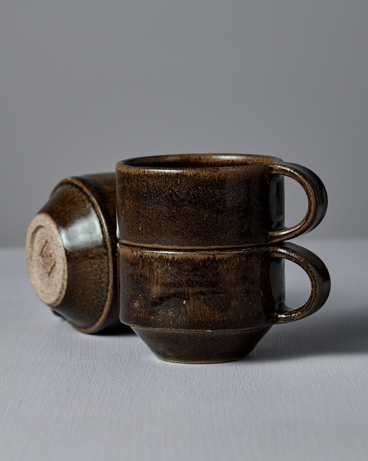 Two Studio Foon Everyday Stacker Cups in Burnt Umber are stacked, with a third cup lying on its side in the background. All are set against a plain grey surface and wall.