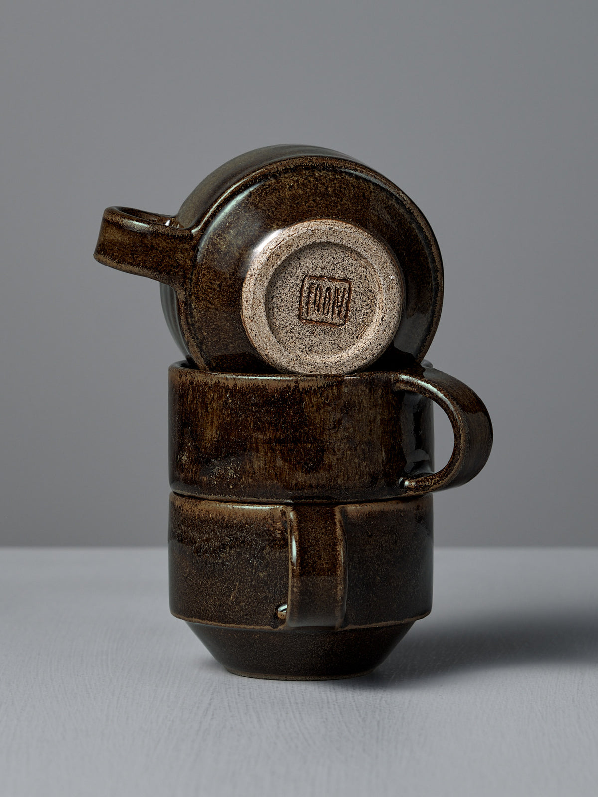 Two stacked Studio Foon Everyday Stacker Cups – Small in Burnt Umber, with the top cup inverted to reveal its stamped maker&#39;s mark, are shown against a plain gray background.