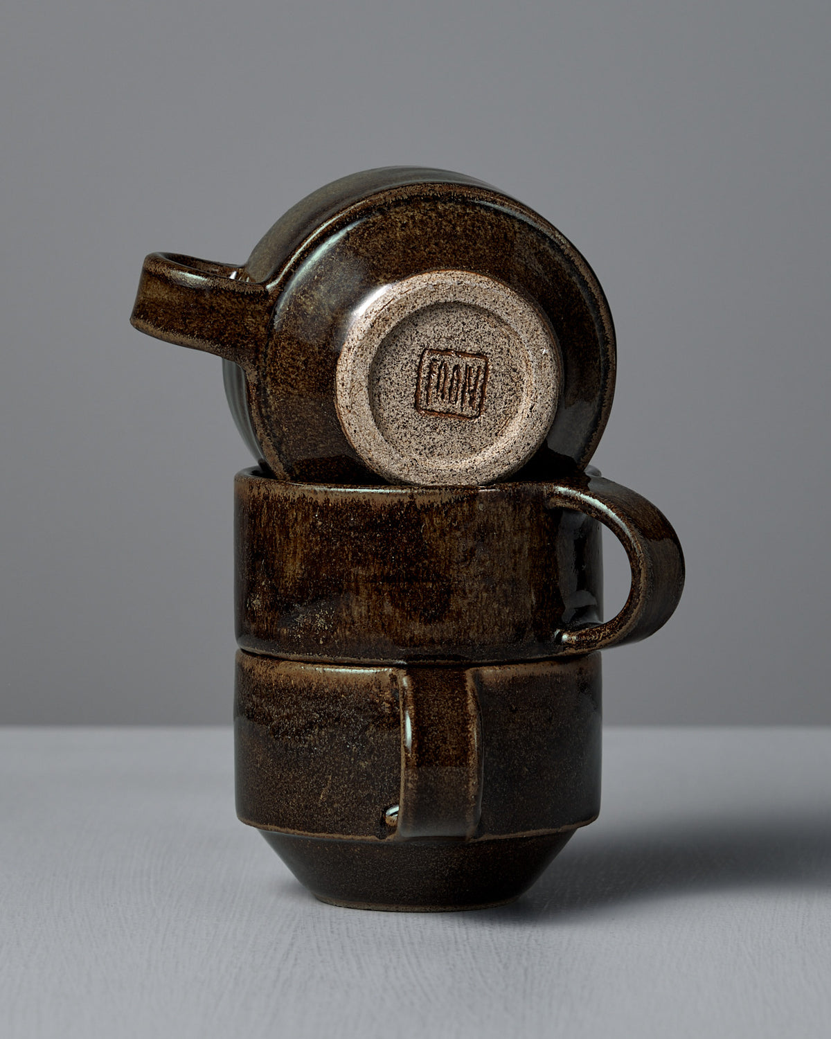Two stacked Studio Foon Everyday Stacker Cups – Small in Burnt Umber, with the top cup inverted to reveal its stamped maker's mark, are shown against a plain gray background.