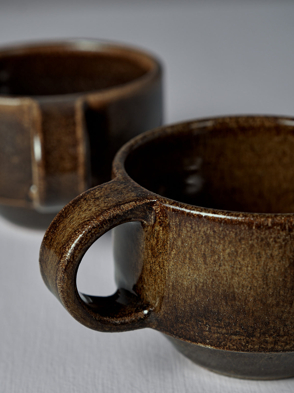 Two Everyday Stacker Cups – Small | Burnt Umber, featuring visible handles and a handcrafted dark brown finish, are placed on a light surface, with one in the foreground and another slightly blurred in the background.