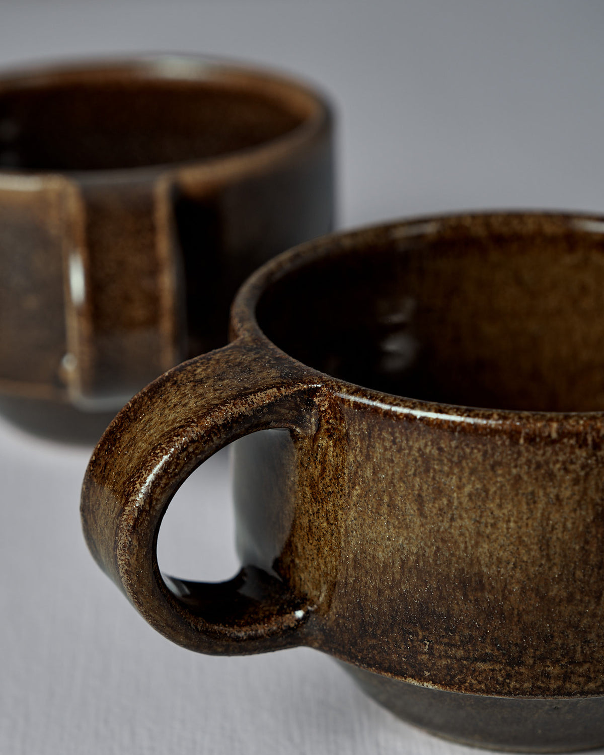 Two Everyday Stacker Cups – Small | Burnt Umber, featuring visible handles and a handcrafted dark brown finish, are placed on a light surface, with one in the foreground and another slightly blurred in the background.