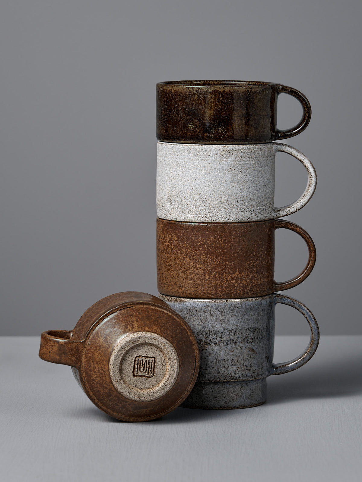 Four Everyday Stacker Cups – Small in Stone White by Studio Foon are stacked on a gray surface, with one cup lying on its side to display the maker’s mark.