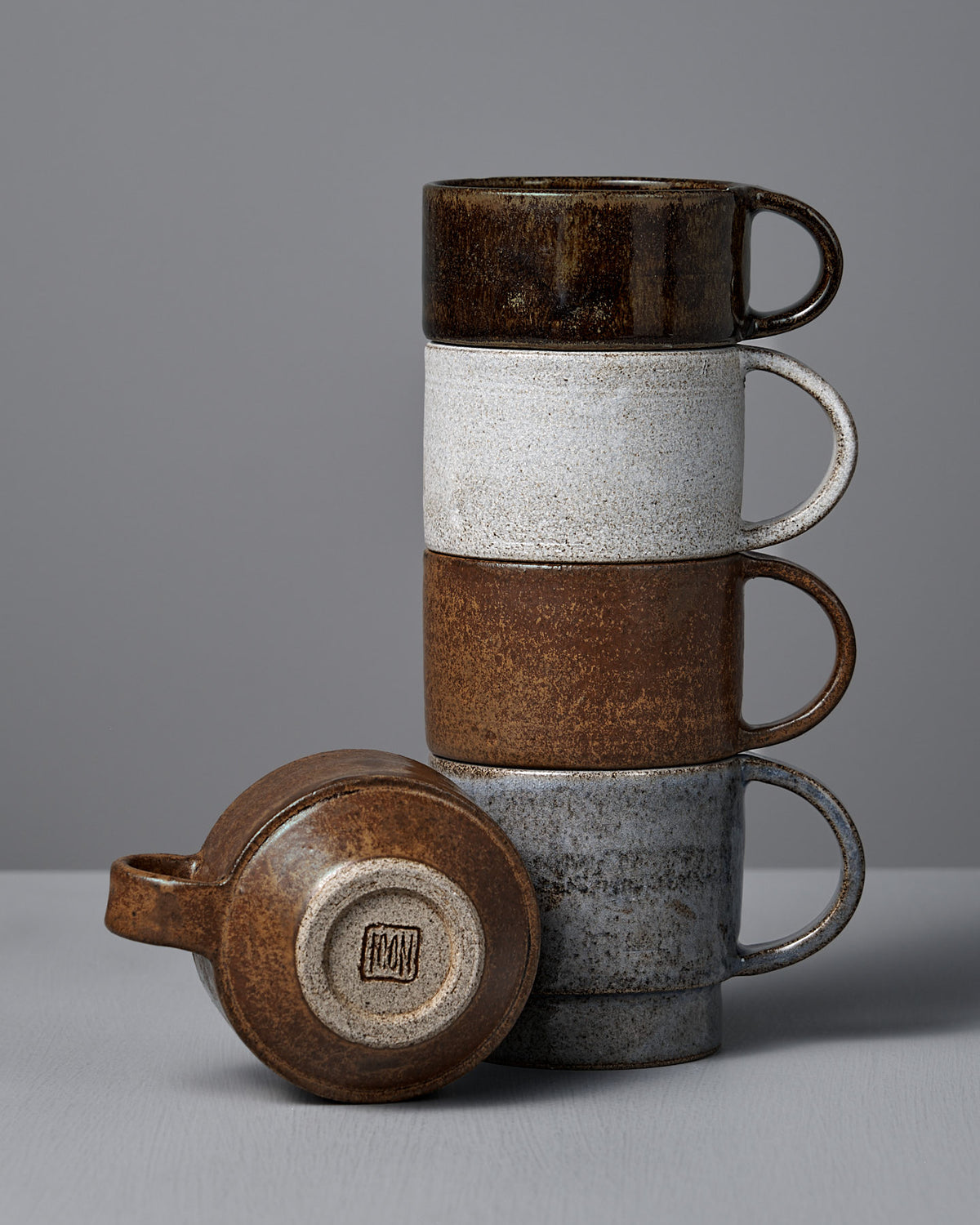 Four Everyday Stacker Cups – Small in Stone White by Studio Foon are stacked on a gray surface, with one cup lying on its side to display the maker’s mark.