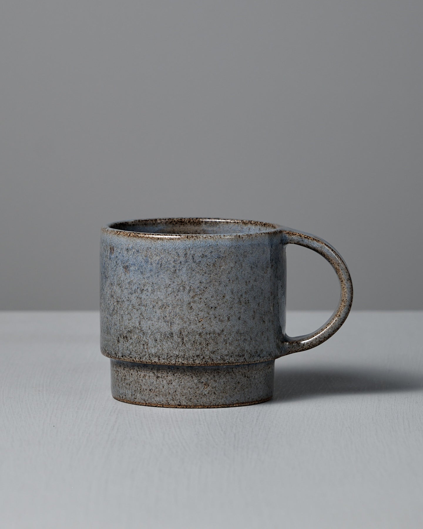 The Studio Foon Everyday Stacker Cup – Large | Indigo, a handcrafted 300ml cup with a speckled gray glaze and rounded handle, rests on a light gray surface against a plain gray background.