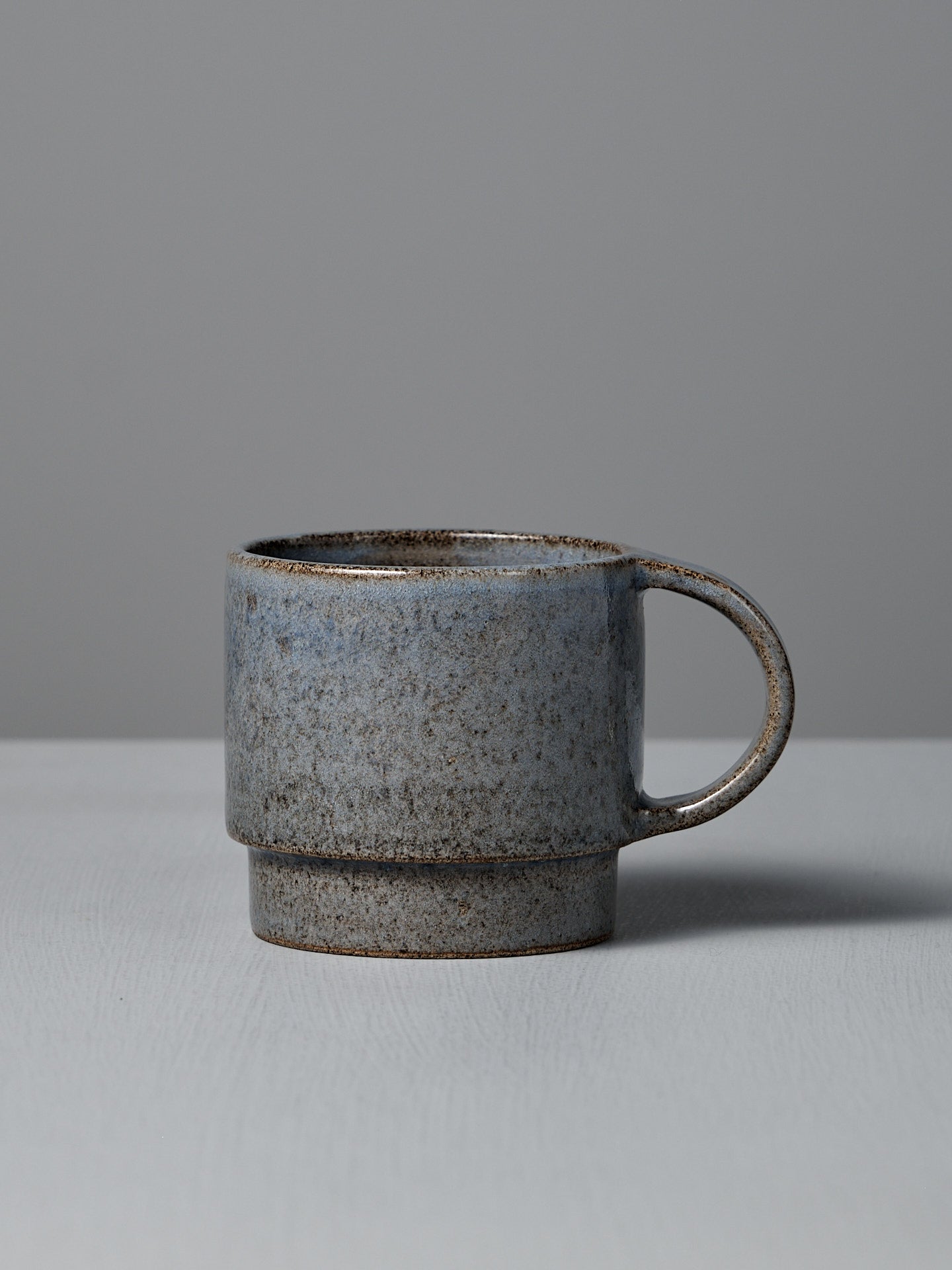 The Studio Foon Everyday Stacker Cup – Large | Indigo, a handcrafted 300ml cup with a speckled gray glaze and rounded handle, rests on a light gray surface against a plain gray background.