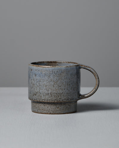 The Studio Foon Everyday Stacker Cup – Large | Indigo, a handcrafted 300ml cup with a speckled gray glaze and rounded handle, rests on a light gray surface against a plain gray background.