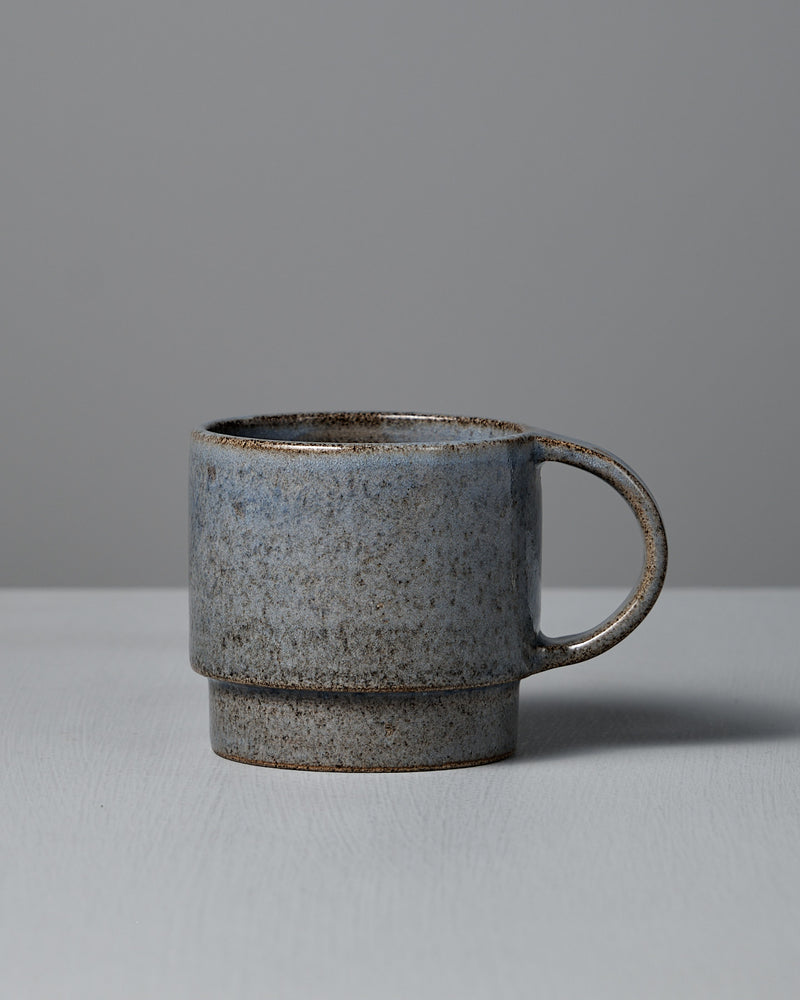The Studio Foon Everyday Stacker Cup – Large | Indigo, a handcrafted 300ml cup with a speckled gray glaze and rounded handle, rests on a light gray surface against a plain gray background.