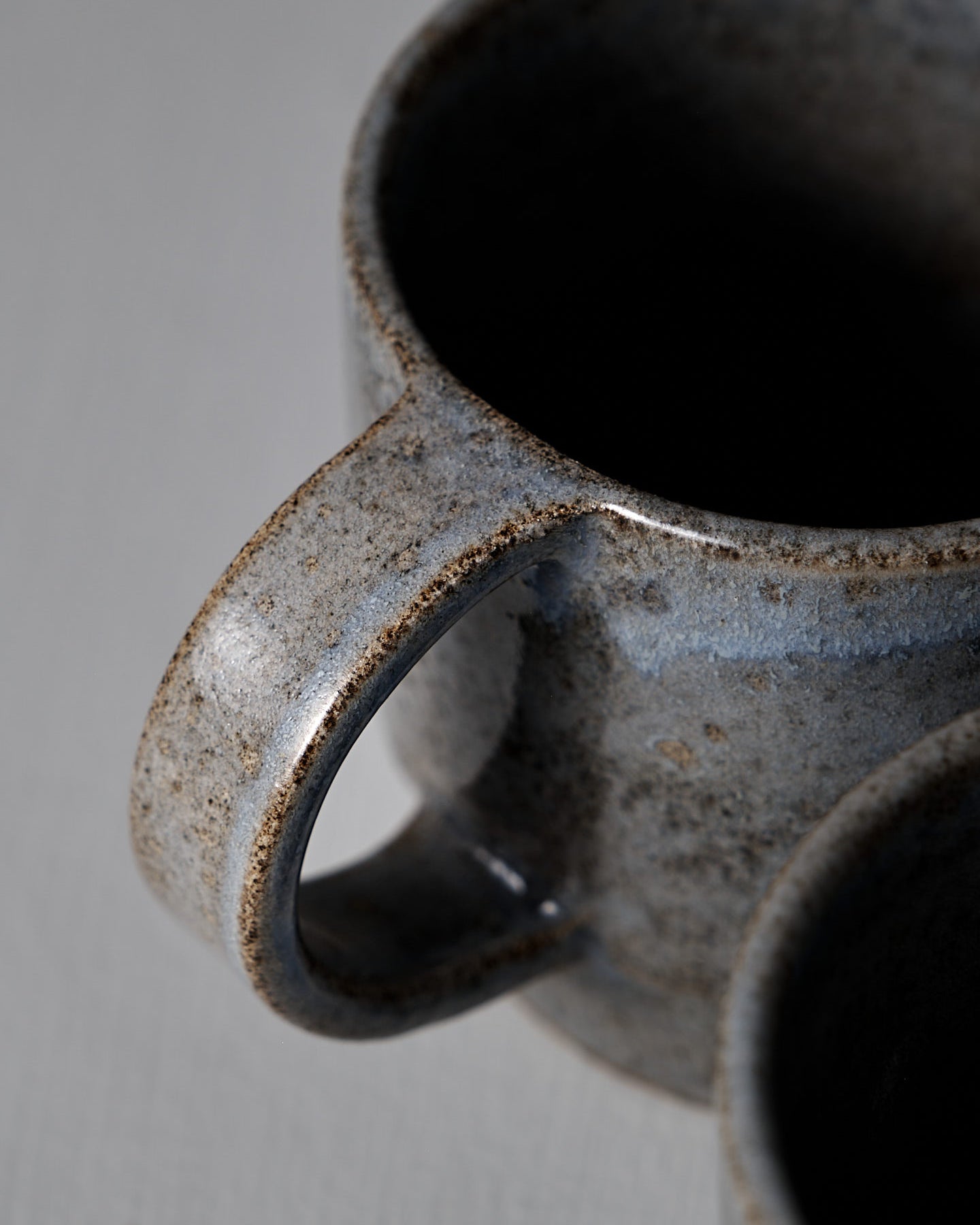 The Studio Foon Everyday Stacker Cup – Large | Indigo, a handcrafted 300ml cup with a speckled gray glaze and rounded handle, rests on a light gray surface against a plain gray background.