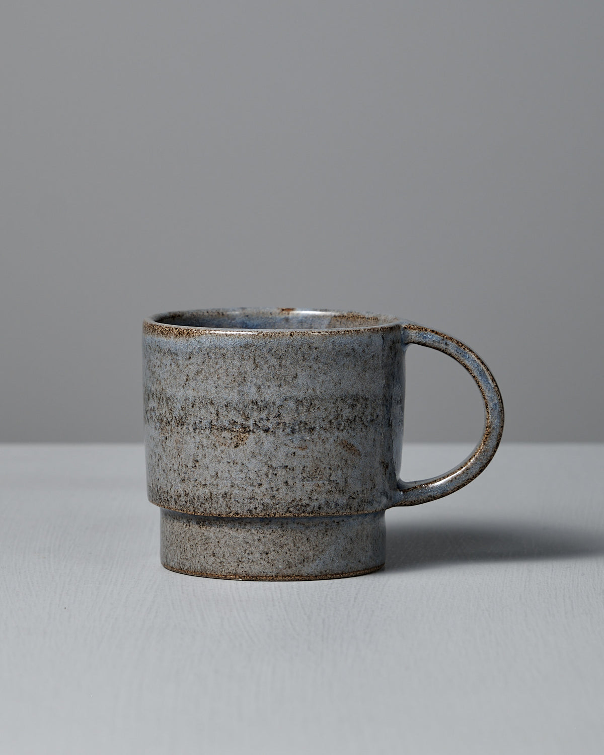 The Studio Foon Everyday Stacker Cup – Large | Indigo (300ml), handcrafted with a speckled gray and brown finish, is displayed on a light surface against a plain gray background.