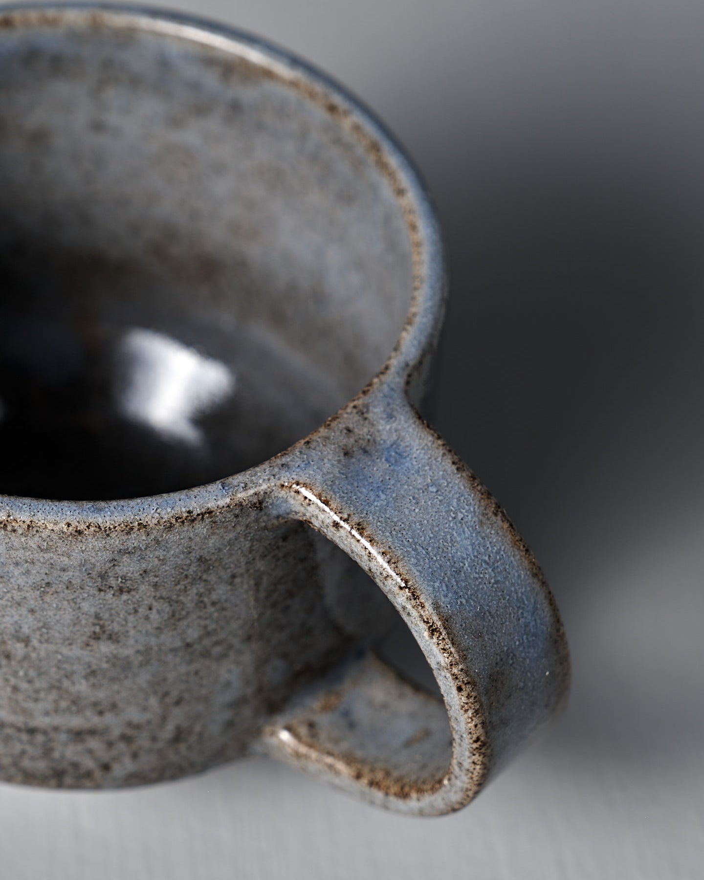 The Studio Foon Everyday Stacker Cup – Medium | Indigo, a handcrafted ceramic cup with handle and speckled gray glaze, rests on a light gray surface against a plain gray background.