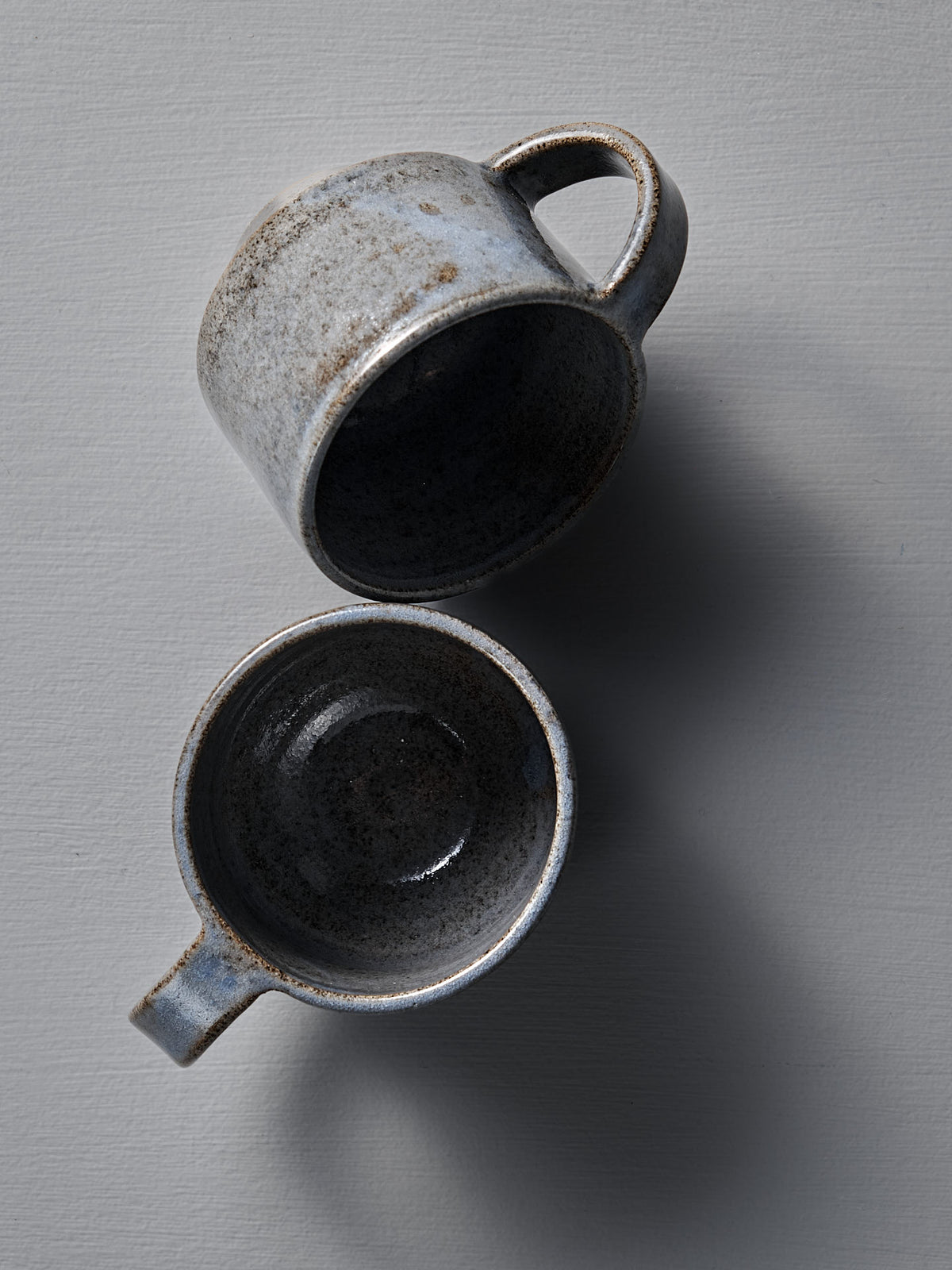Two indigo Everyday Stacker Cups – Medium by Studio Foon with handles are displayed on a light gray surface, one upright and the other lying on its side.