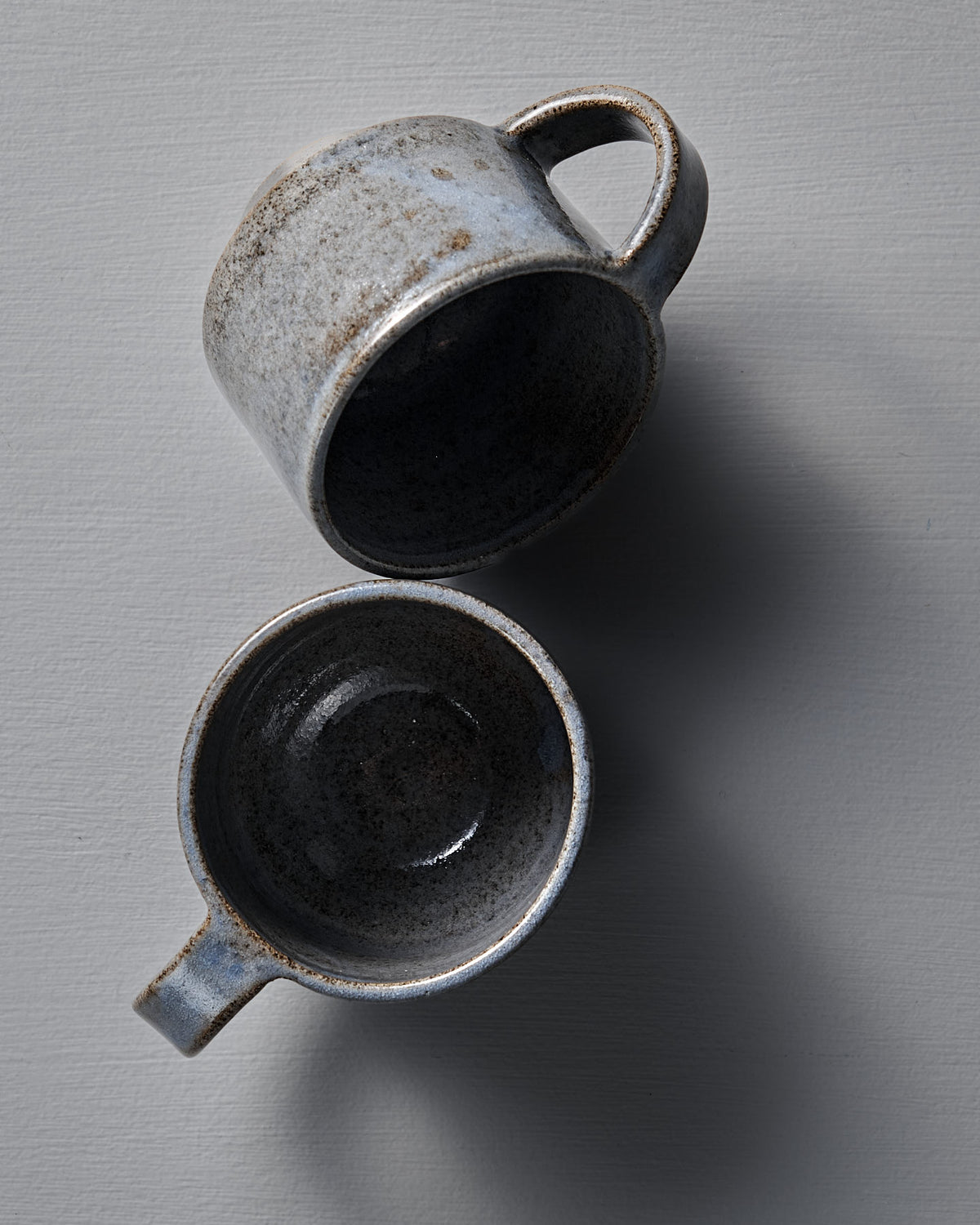 Two indigo Everyday Stacker Cups – Medium by Studio Foon with handles are displayed on a light gray surface, one upright and the other lying on its side.