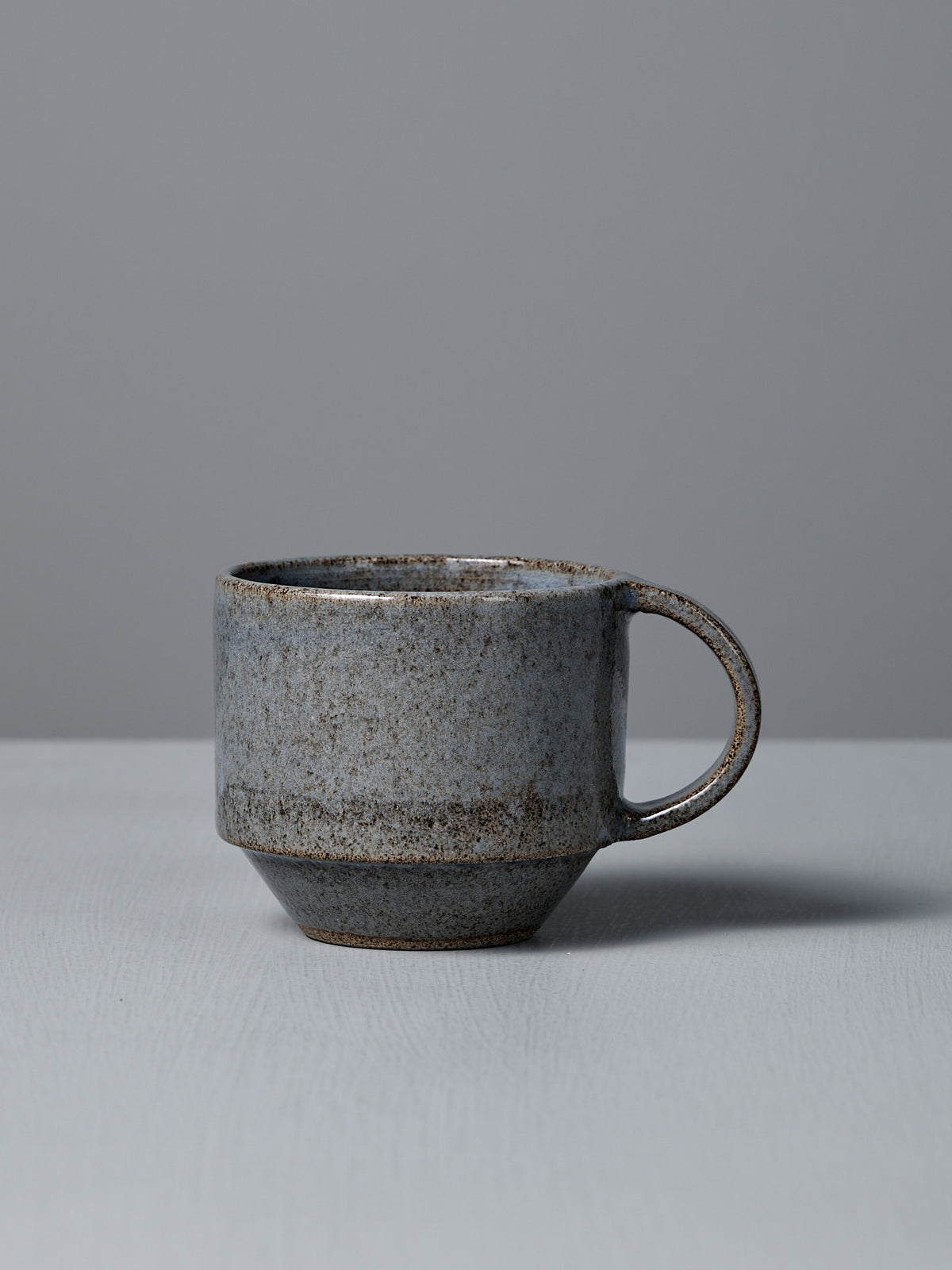 The Studio Foon Everyday Stacker Cup – Medium | Indigo, a handcrafted ceramic cup with handle and speckled gray glaze, rests on a light gray surface against a plain gray background.