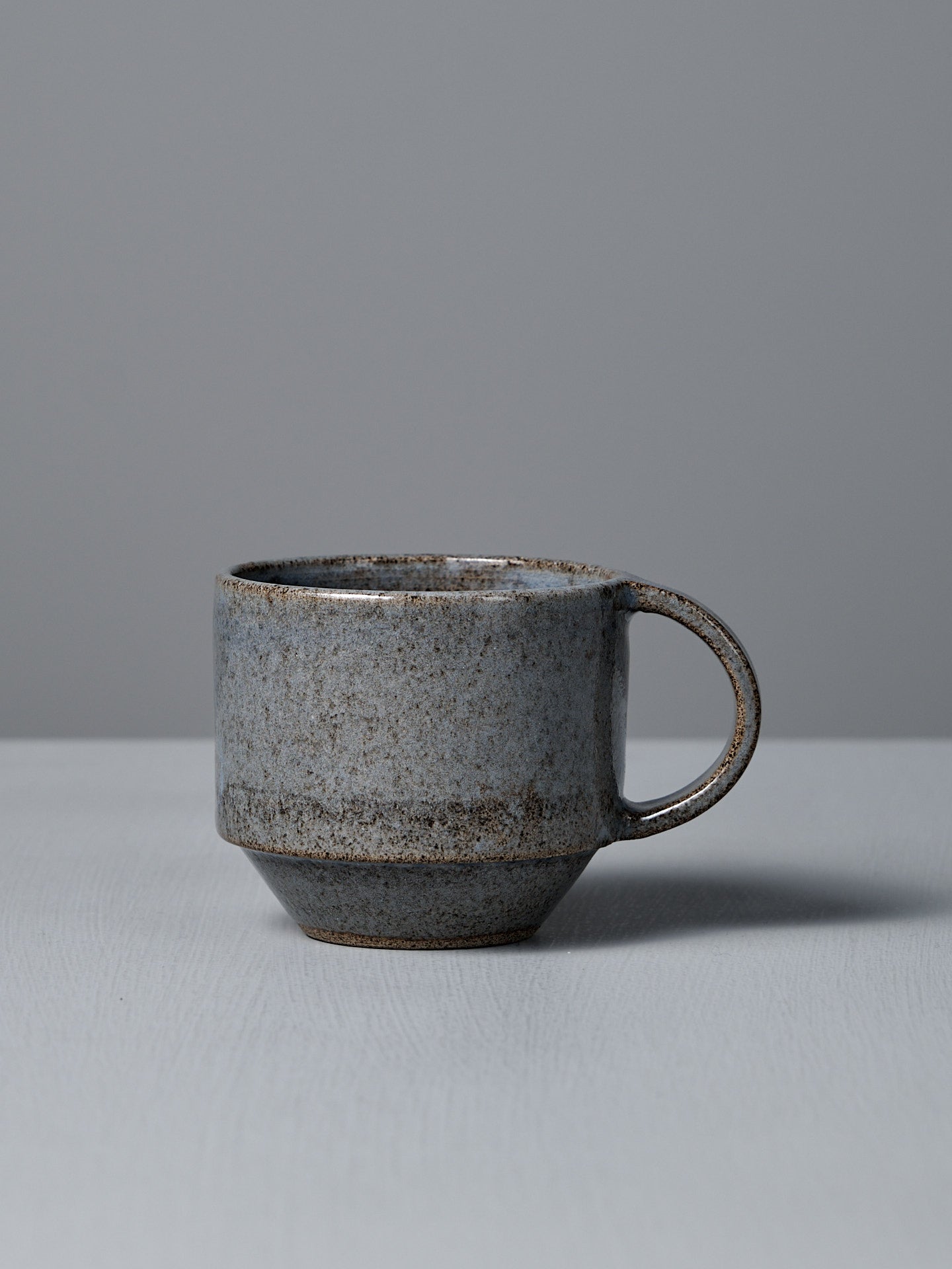 The Studio Foon Everyday Stacker Cup – Medium | Indigo, a handcrafted ceramic cup with handle and speckled gray glaze, rests on a light gray surface against a plain gray background.