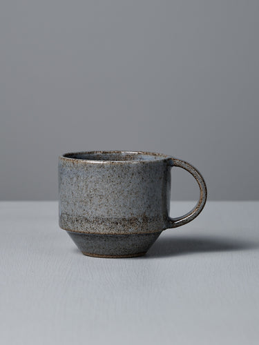 The Studio Foon Everyday Stacker Cup – Medium | Indigo, a handcrafted ceramic cup with handle and speckled gray glaze, rests on a light gray surface against a plain gray background.