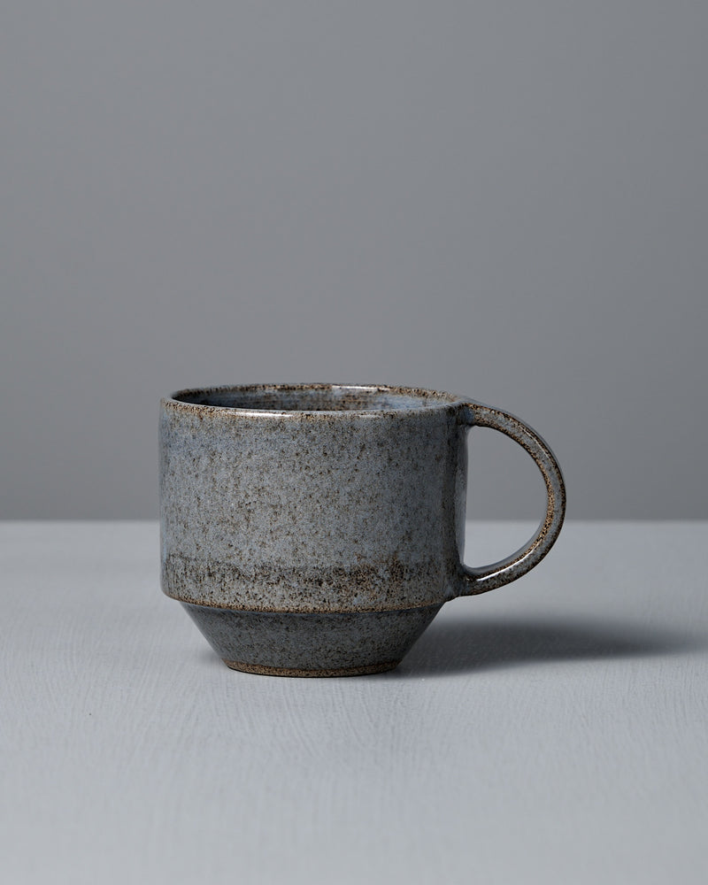 The Studio Foon Everyday Stacker Cup – Medium | Indigo, a handcrafted ceramic cup with handle and speckled gray glaze, rests on a light gray surface against a plain gray background.