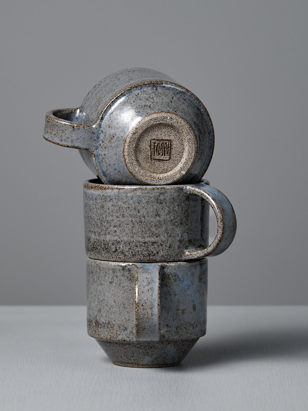 The Studio Foon Everyday Stacker Cup – Medium in Indigo features a handcrafted, rough speckled texture and muted blue-grey tones, shown here as three stacked cups against a plain gray background.