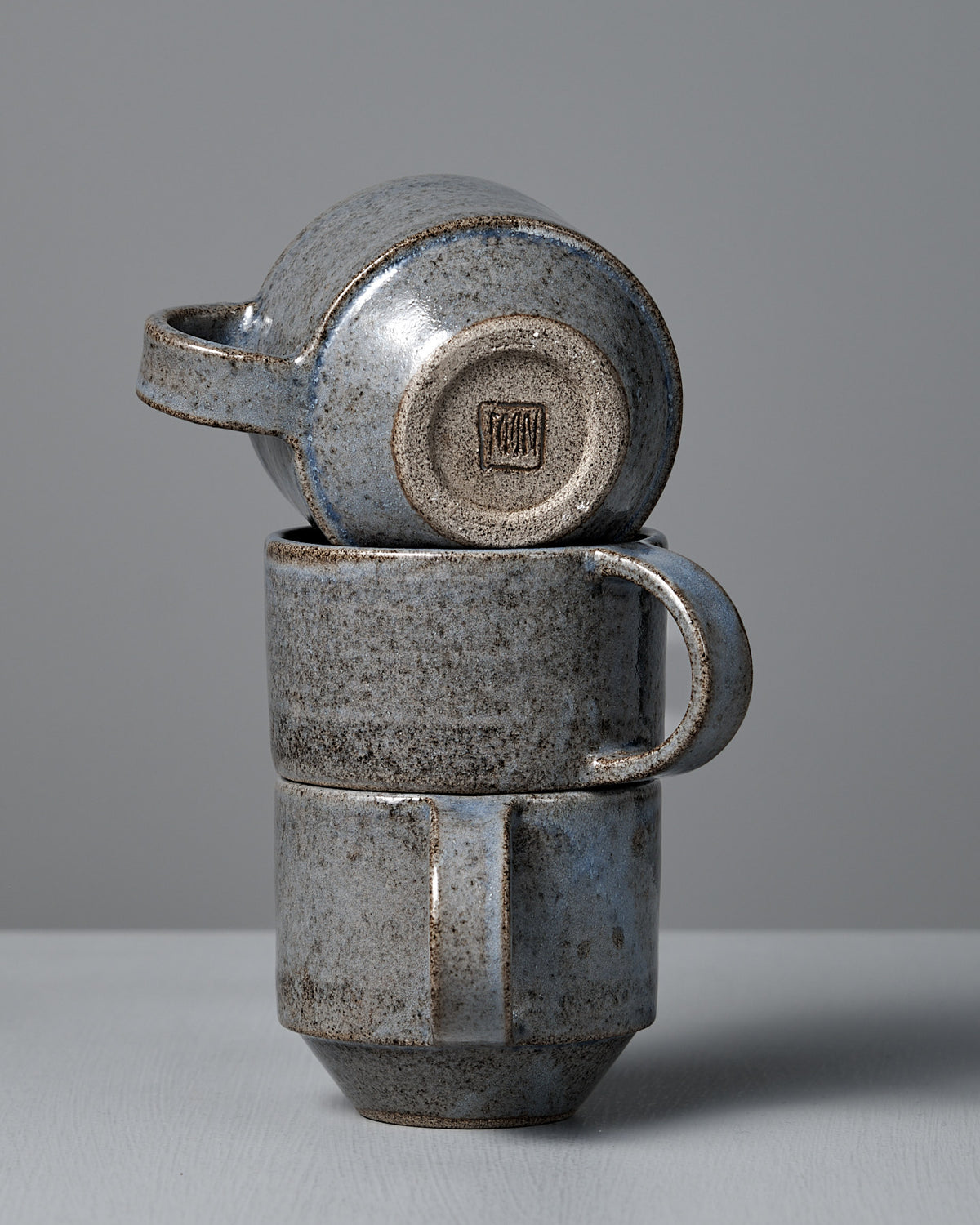 The Studio Foon Everyday Stacker Cup – Medium in Indigo features a handcrafted, rough speckled texture and muted blue-grey tones, shown here as three stacked cups against a plain gray background.
