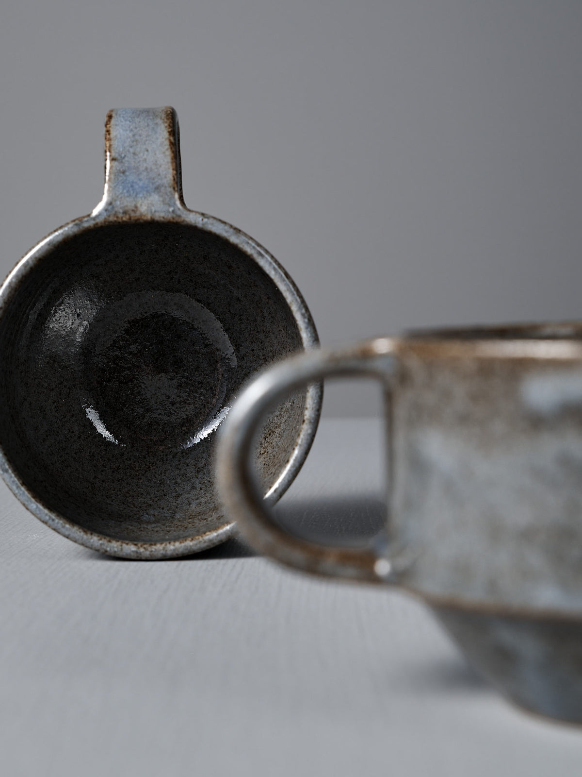 Two Studio Foon Everyday Stacker Cups – Small | Indigo, featuring a blue-gray speckled glaze, rest on a light gray surface—one upright and one on its side to reveal the interior.