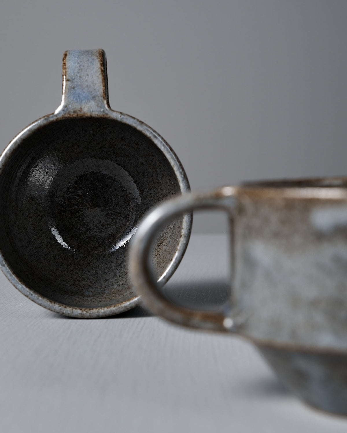 Two Studio Foon Everyday Stacker Cups – Small | Indigo, featuring a blue-gray speckled glaze, rest on a light gray surface—one upright and one on its side to reveal the interior.