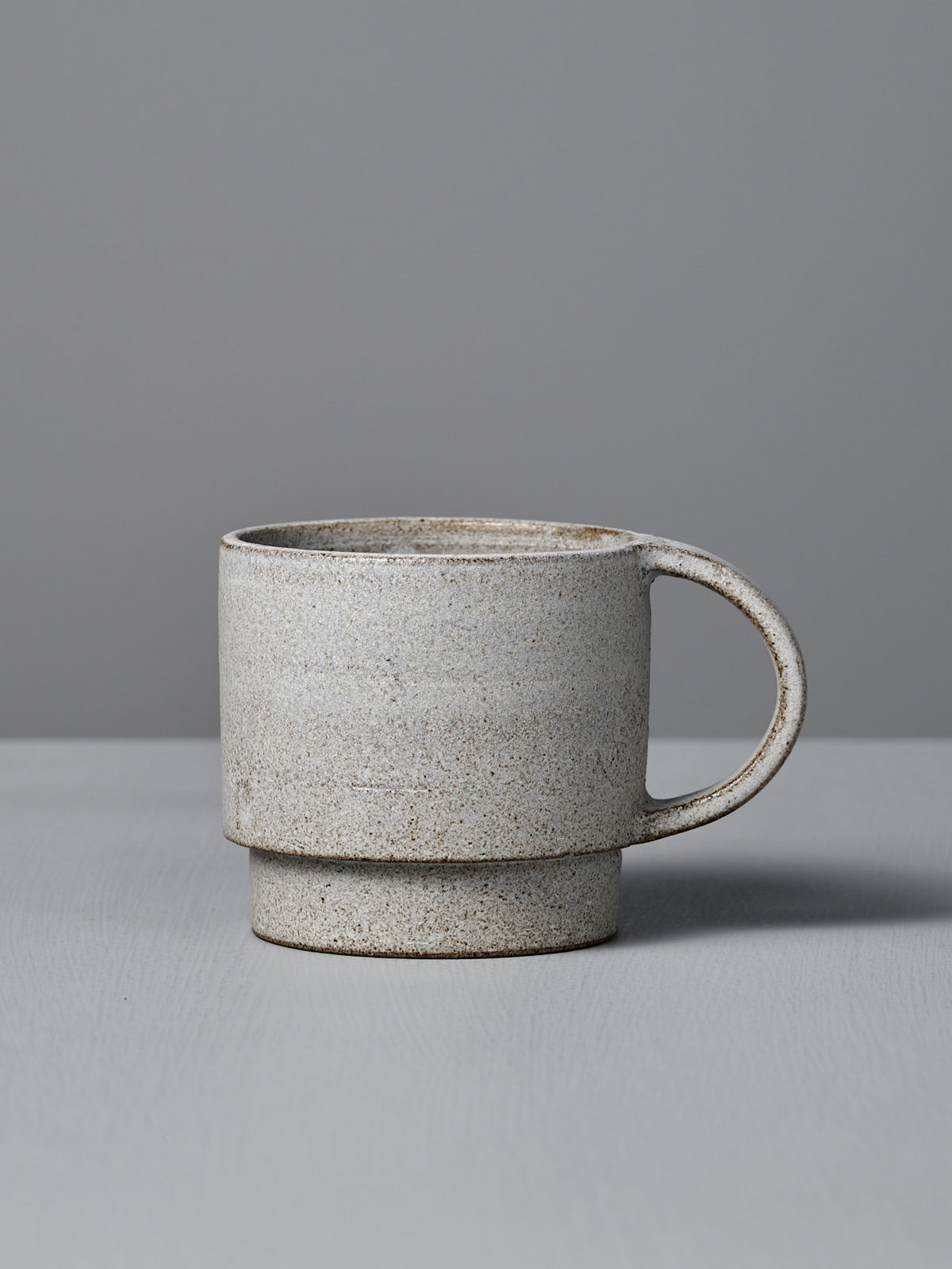 The Studio Foon Everyday Stacker Cup – Large | Stone White is a handcrafted, speckled 300ml cup with a large handle. It’s perfect for daily use and pairs well with other stackable cups on any light gray surface.