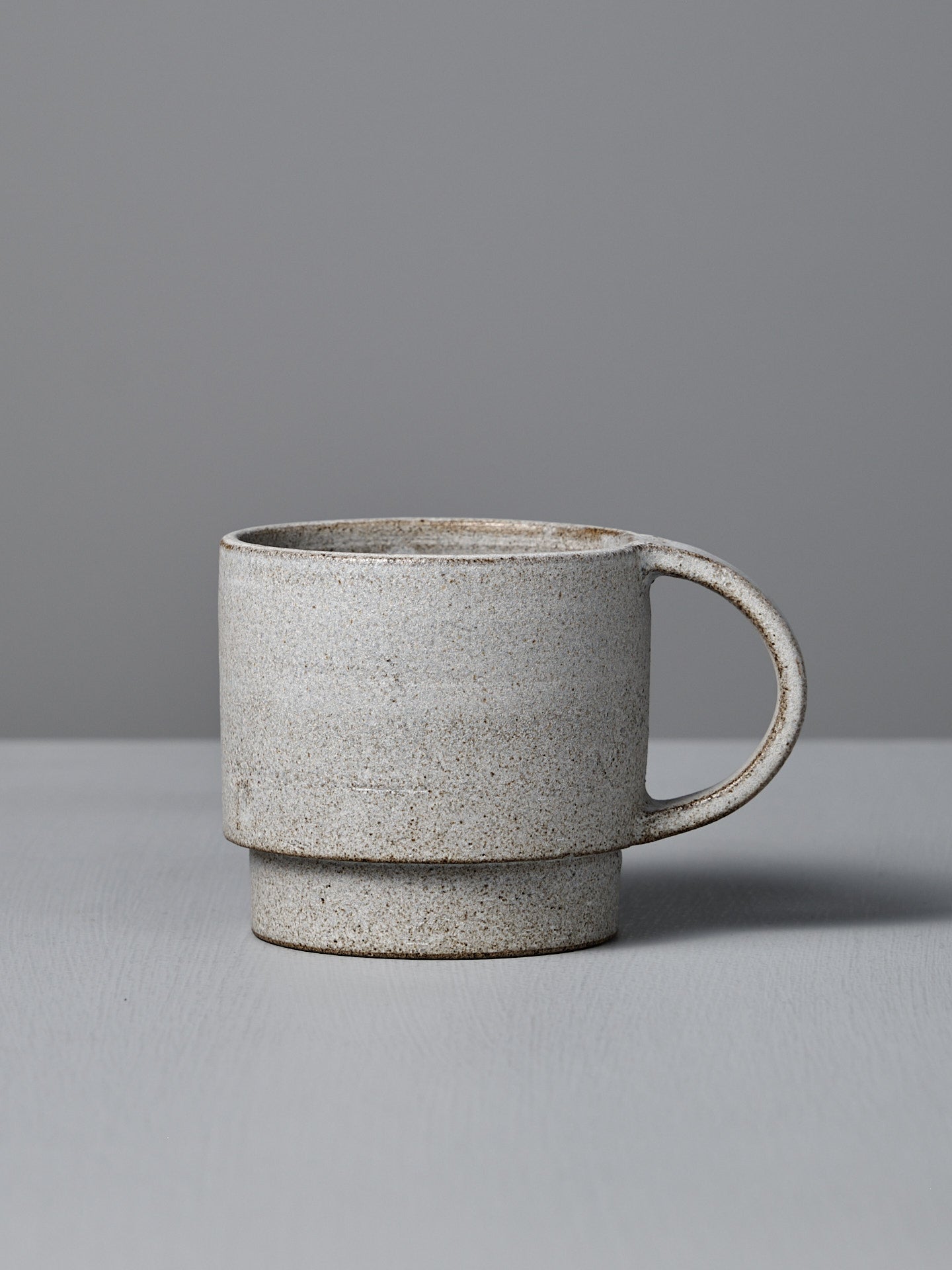 The Studio Foon Everyday Stacker Cup – Large | Stone White is a handcrafted, speckled 300ml cup with a large handle. It’s perfect for daily use and pairs well with other stackable cups on any light gray surface.