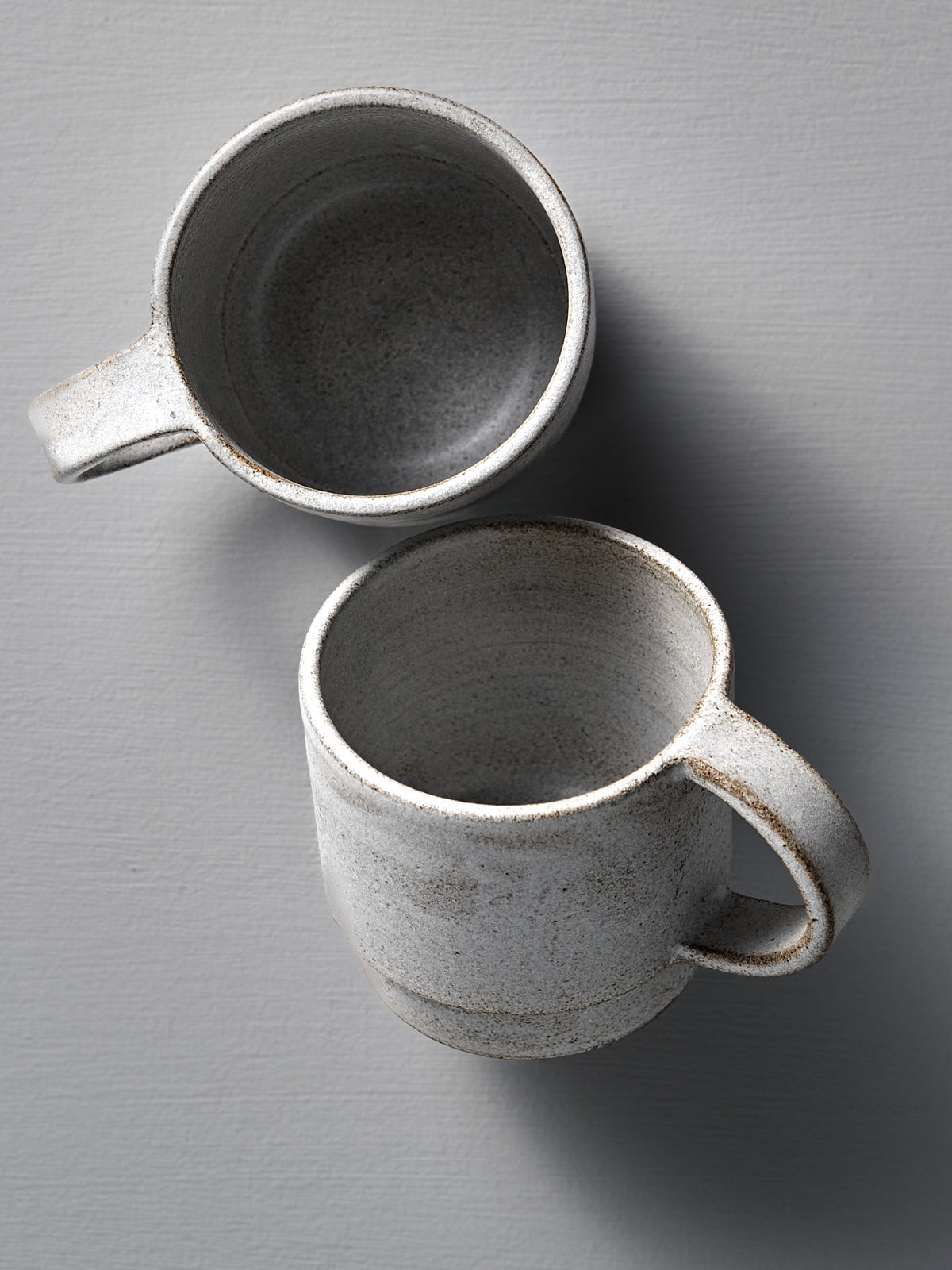 Two Stone White ceramic Everyday Stacker Cups by Studio Foon—one upright, one on its side—rest on a light gray textured surface. Handcrafted and large, they&#39;re perfect for daily use.