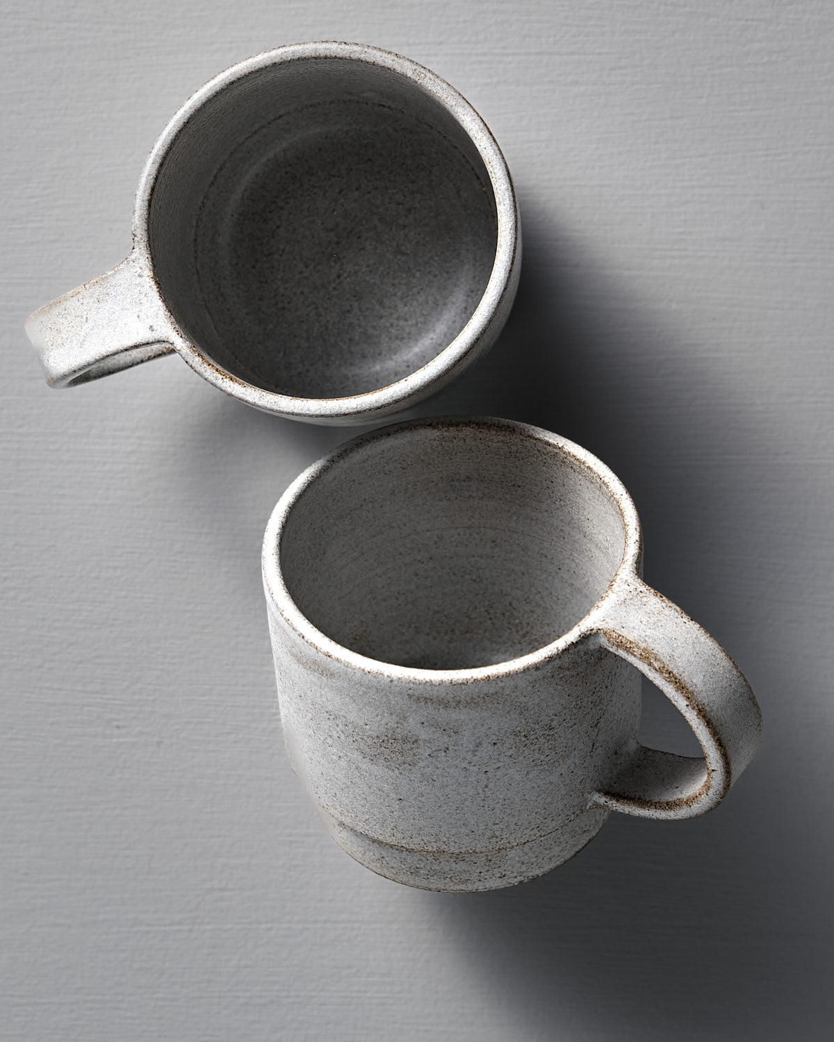 Two Stone White ceramic Everyday Stacker Cups by Studio Foon—one upright, one on its side—rest on a light gray textured surface. Handcrafted and large, they're perfect for daily use.