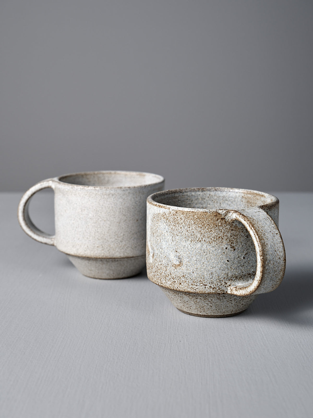 Two Studio Foon Everyday Stacker Cups – Medium in Stone White, featuring large handles and a speckled handcrafted finish, are placed on a light gray surface with a plain gray background.