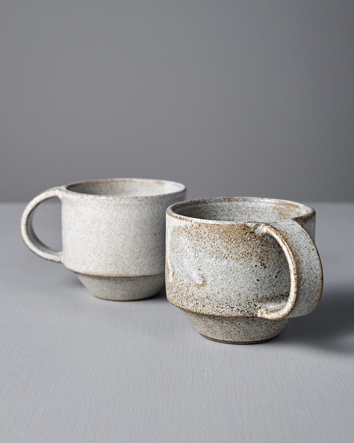 Two Studio Foon Everyday Stacker Cups – Medium in Stone White, featuring large handles and a speckled handcrafted finish, are placed on a light gray surface with a plain gray background.