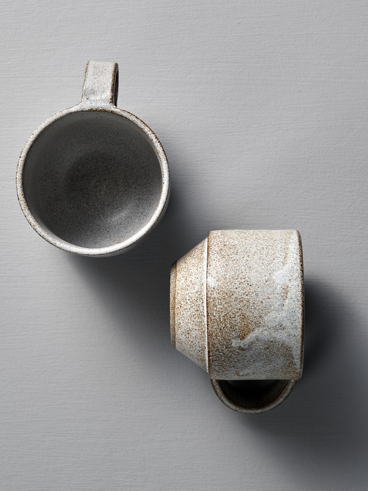 Two handcrafted Studio Foon Everyday Stacker Cups (Medium, Stone White) rest on a light gray surface—one upright, one on its side—ideal for elevating your daily routine.