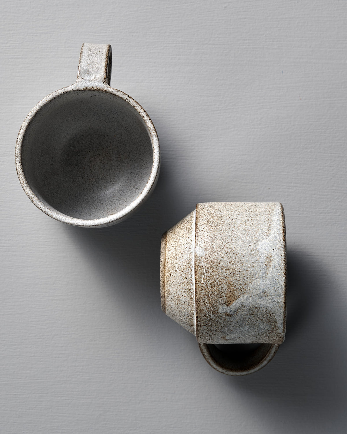 Two handcrafted Studio Foon Everyday Stacker Cups (Medium, Stone White) rest on a light gray surface—one upright, one on its side—ideal for elevating your daily routine.