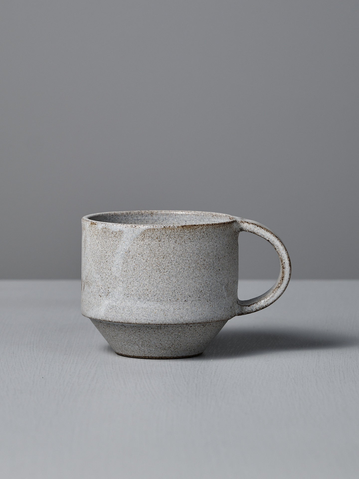 The Studio Foon Everyday Stacker Cup – Medium in Stone White features a matte, speckled finish and sits on a light gray surface, making it perfect for your daily coffee.