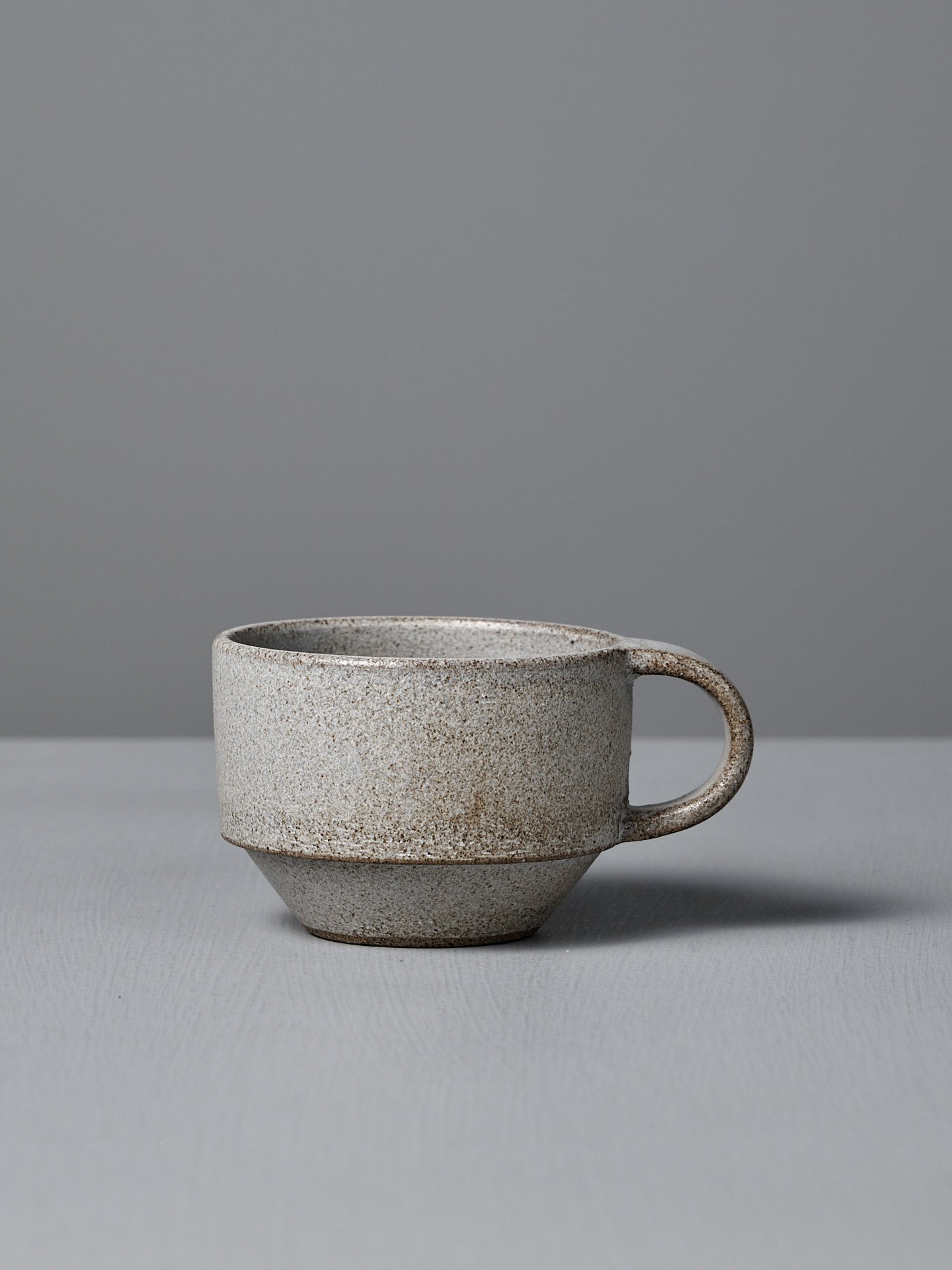 The Studio Foon Everyday Stacker Cup – Small in Stone White features a matte, speckled light gray finish with a handle, resting on a matching surface—ideal for your daily coffee ritual.