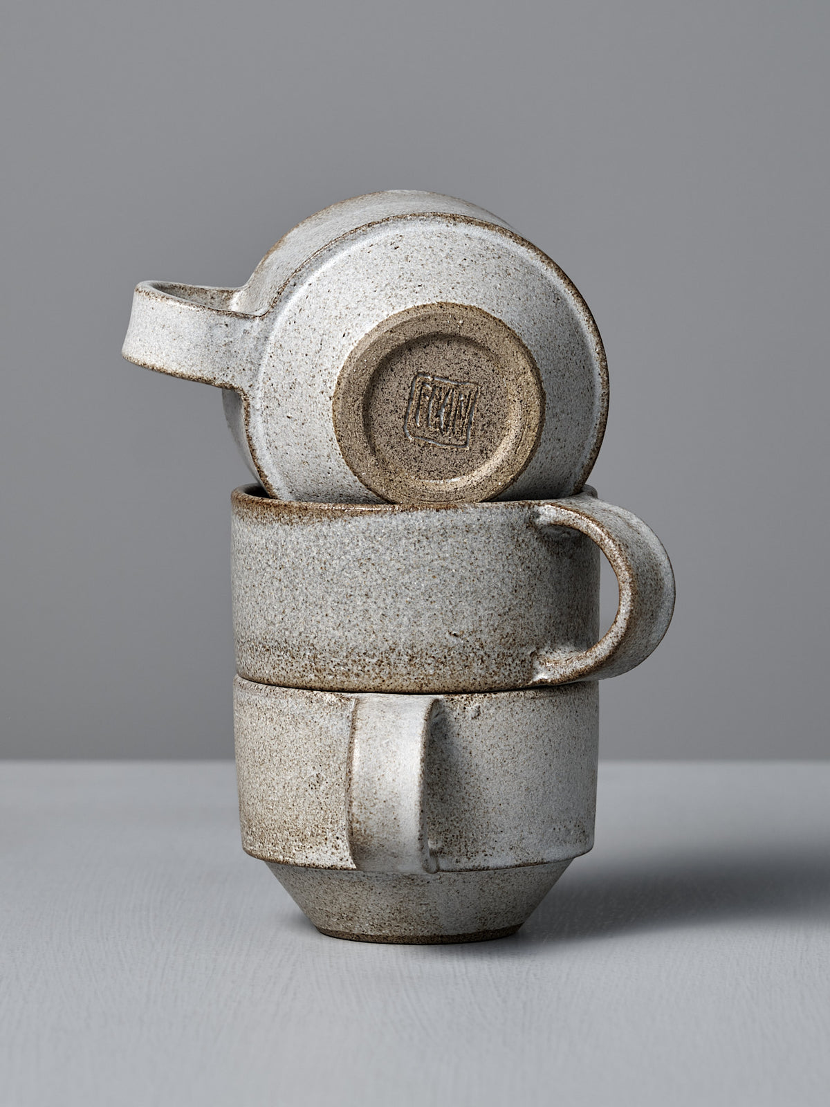 Three Studio Foon Everyday Stacker Cups in Stone White—two upright, one upside down on top—highlight the charm of stackable mugs against a plain gray background.
