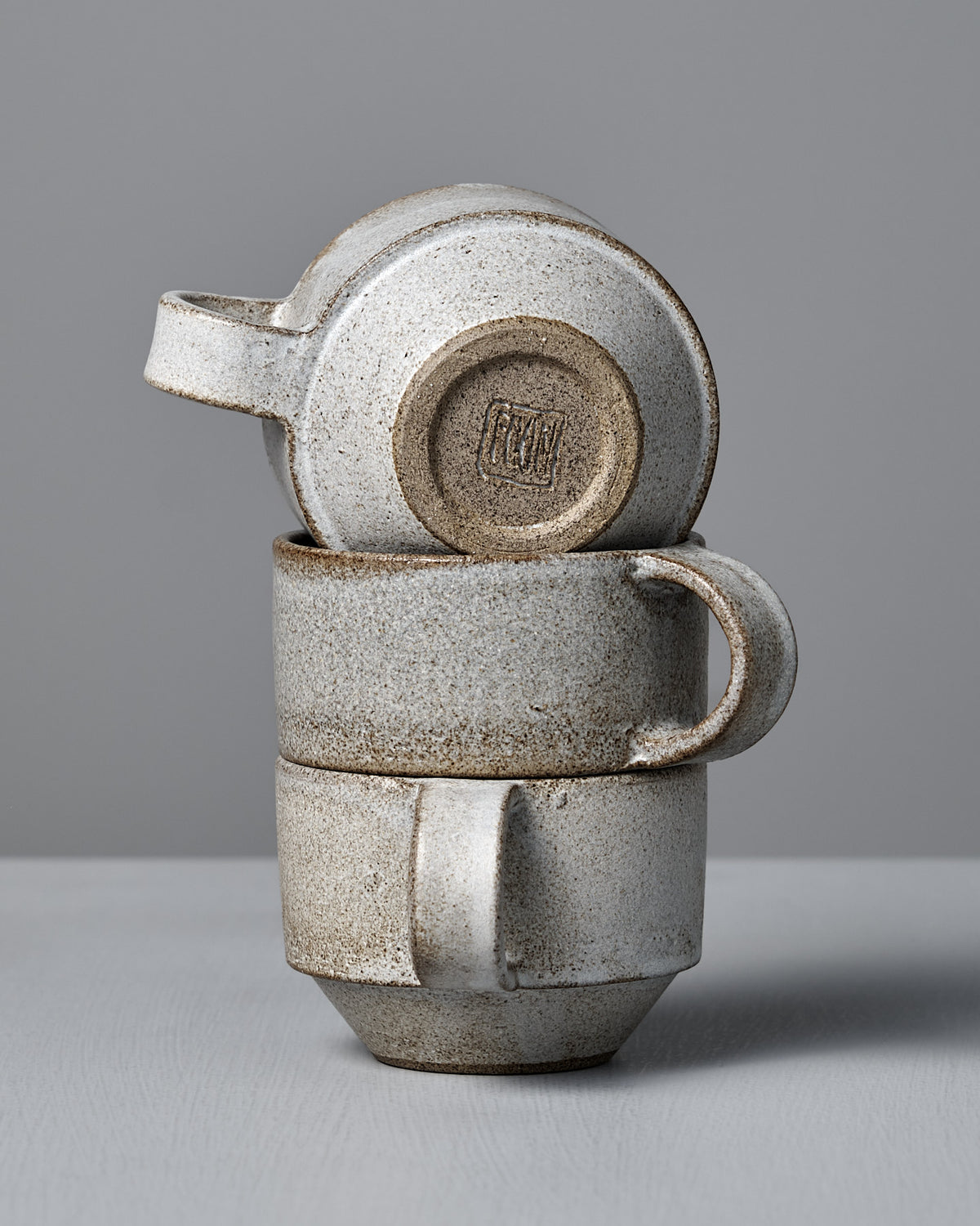 Three Studio Foon Everyday Stacker Cups in Stone White—two upright, one upside down on top—highlight the charm of stackable mugs against a plain gray background.