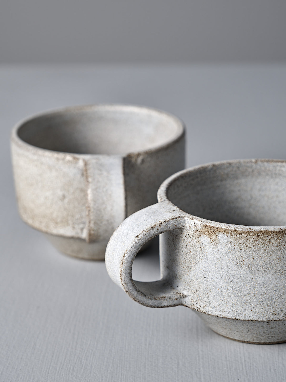 Two Studio Foon Everyday Stacker Cups – Small in Stone White, featuring a handcrafted look and speckled matte finish, sit on a light gray surface against a neutral backdrop.