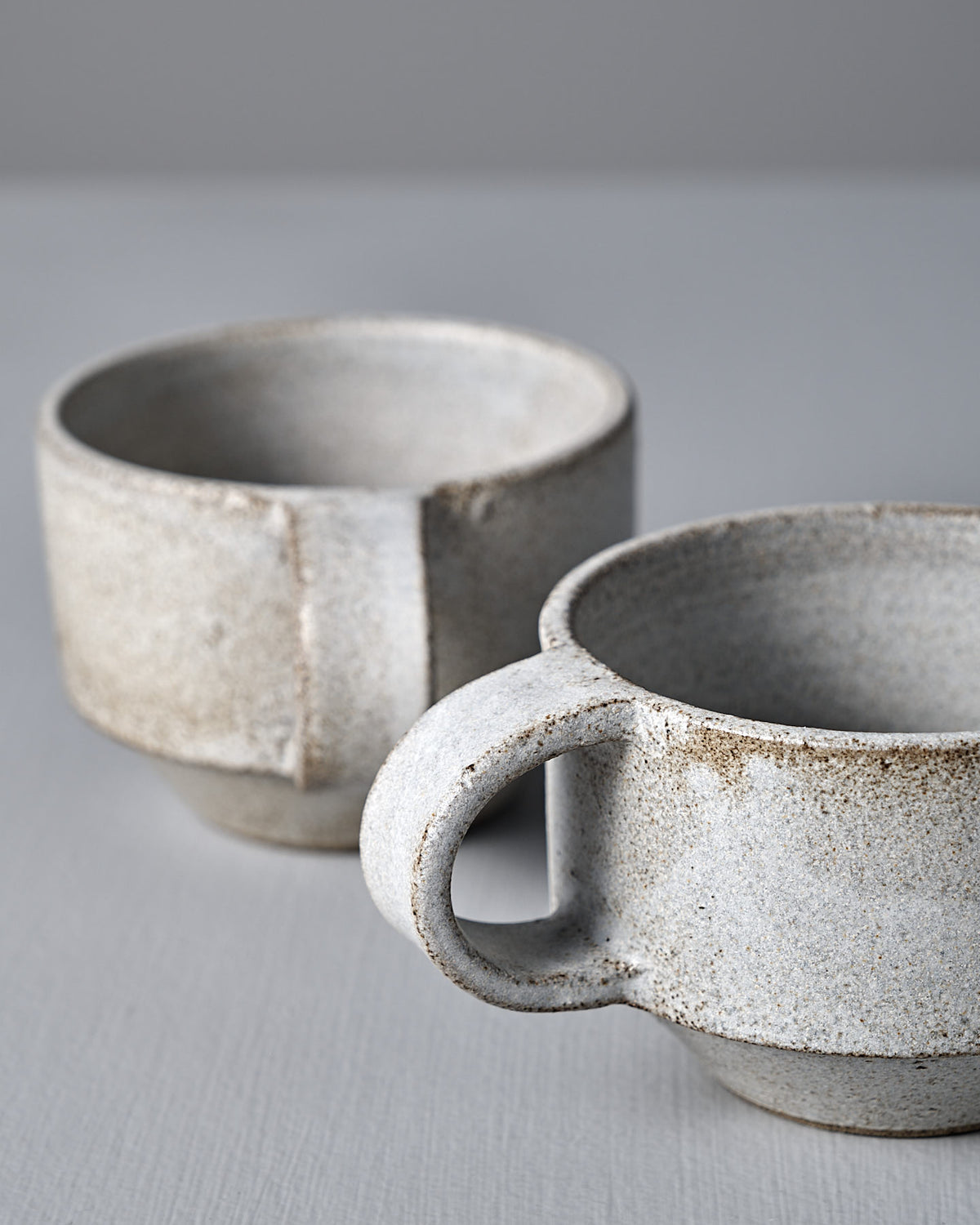 Two Studio Foon Everyday Stacker Cups – Small in Stone White, featuring a handcrafted look and speckled matte finish, sit on a light gray surface against a neutral backdrop.