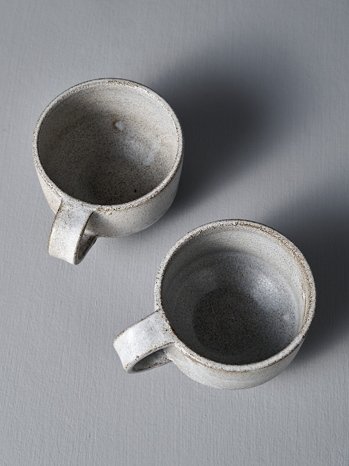 Two empty Studio Foon Everyday Stacker Cups – Small in Stone White, each with a handle, are set at a slight angle to one another on a gray textured surface.