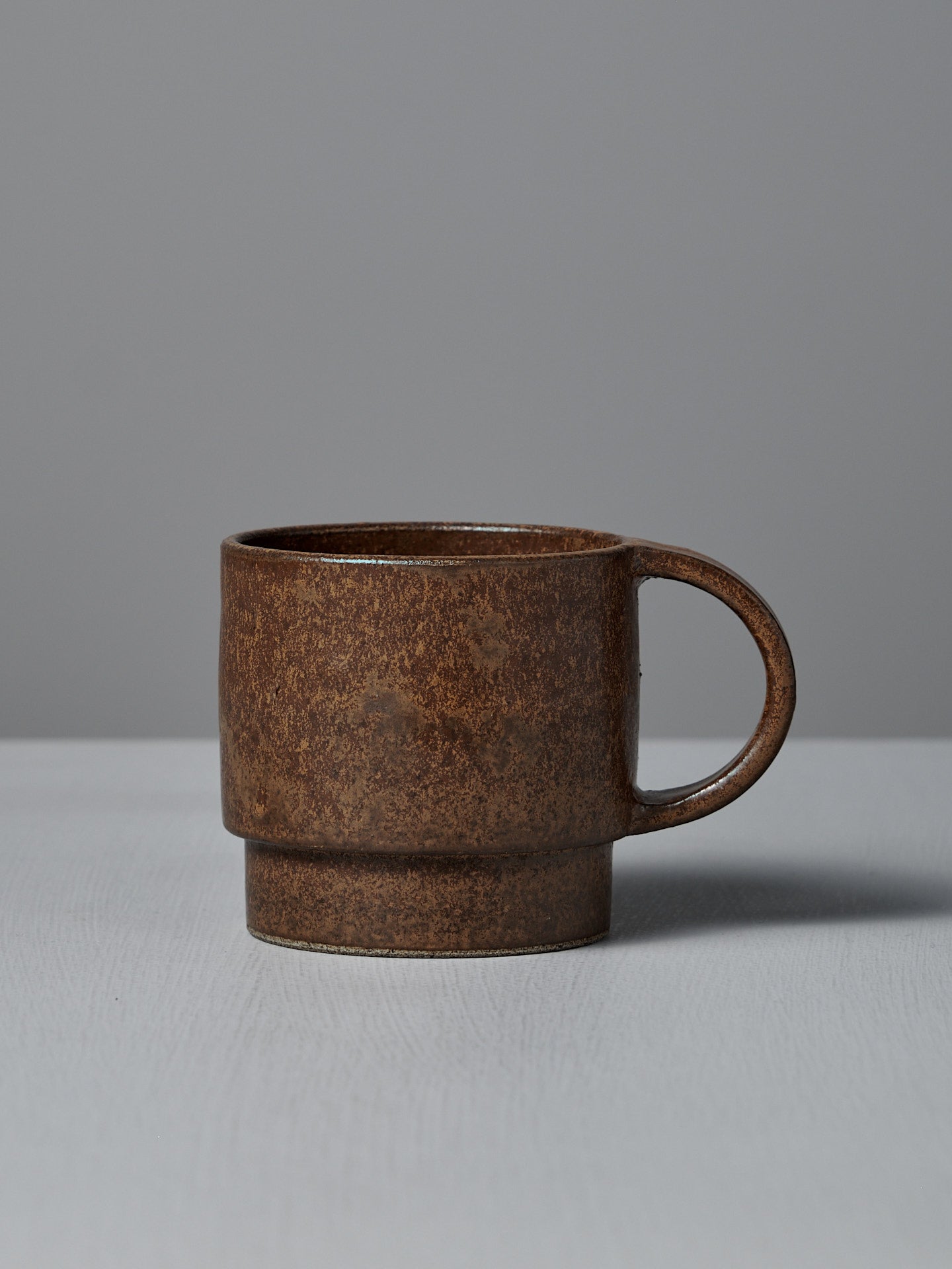 The Studio Foon Everyday Stacker Cup – Large in terracotta, featuring a rounded handle, is ideal for daily collections and is displayed on a light gray surface against a plain gray background.