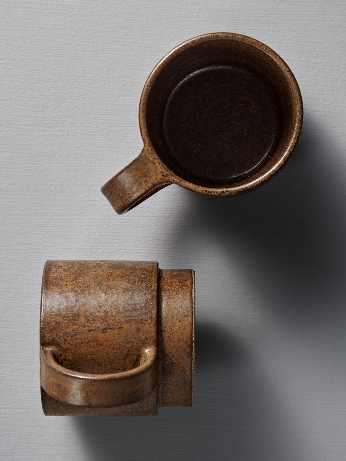 Two Studio Foon Everyday Stacker Cups – Large in terracotta: one viewed from above, partially filled with a dark liquid, the other from the side. Both rest on a light gray textured surface.