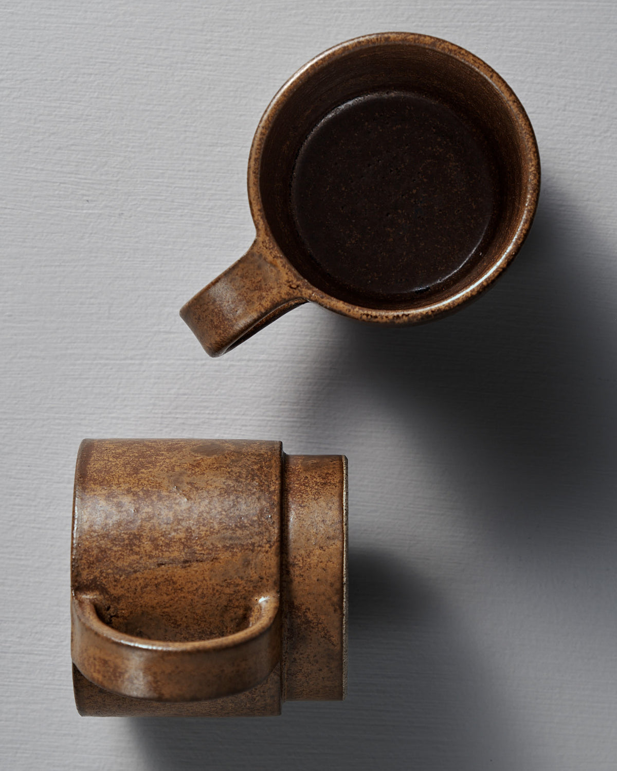 Two Studio Foon Everyday Stacker Cups – Large in terracotta: one viewed from above, partially filled with a dark liquid, the other from the side. Both rest on a light gray textured surface.