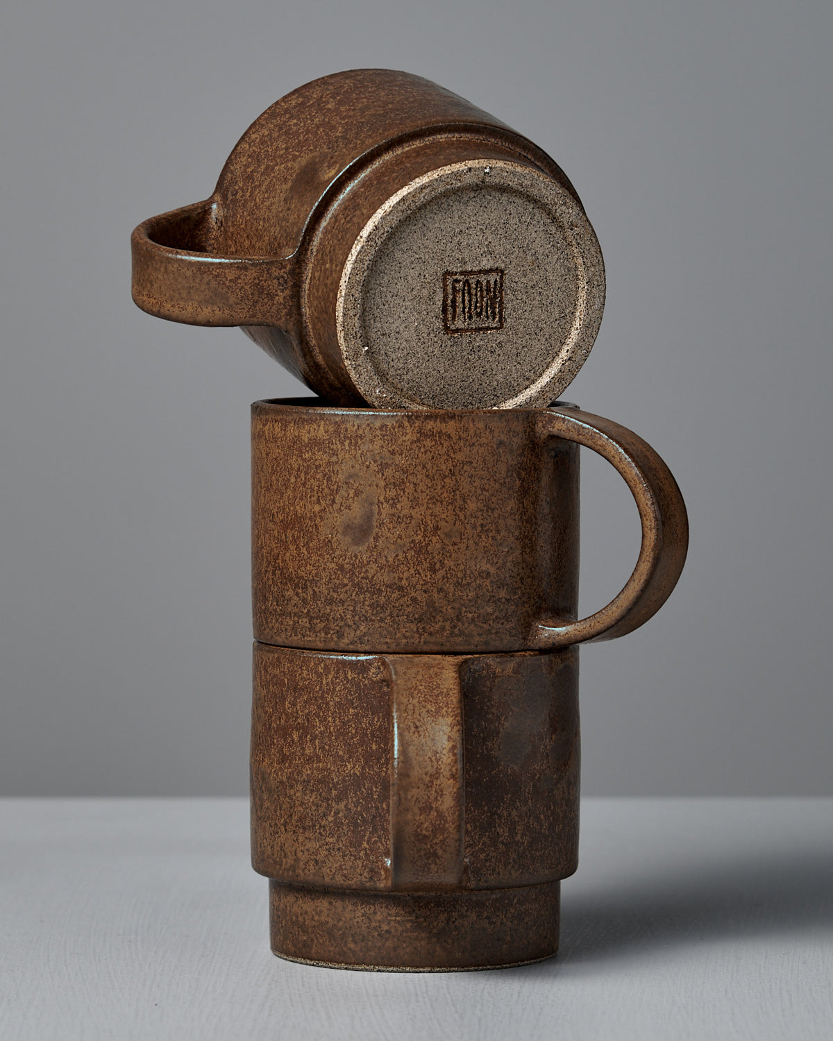 Three Studio Foon Everyday Stacker Cups – Large in terracotta are arranged with two stacked and a third resting sideways on top, all set against a plain gray background.