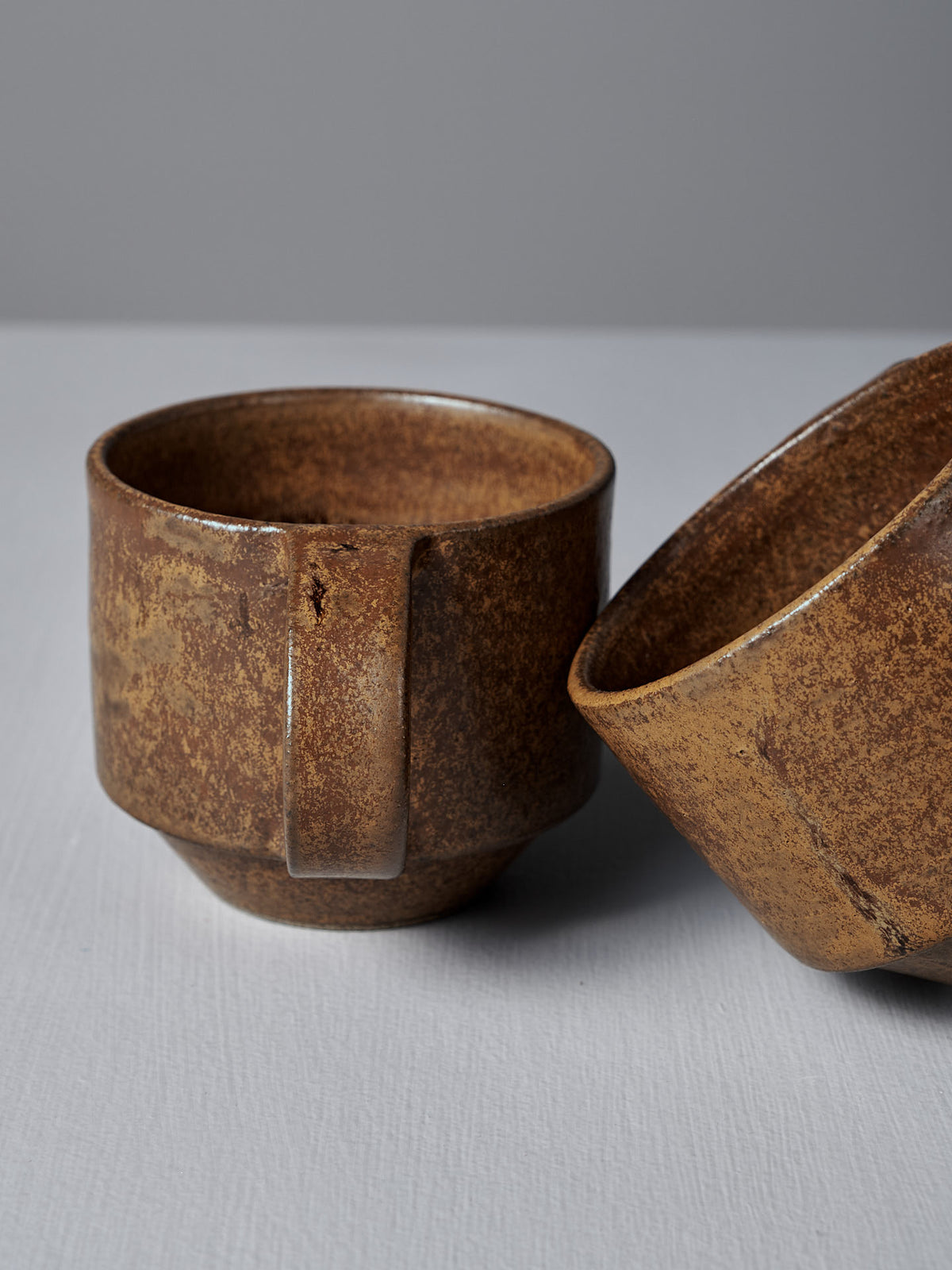 Two Studio Foon Everyday Stacker Cups – Medium in terracotta, with a rustic, textured finish, are set on a light gray surface; one stands upright while the other lies on its side. Both are dishwasher safe and ideal for a handcrafted cup collection.