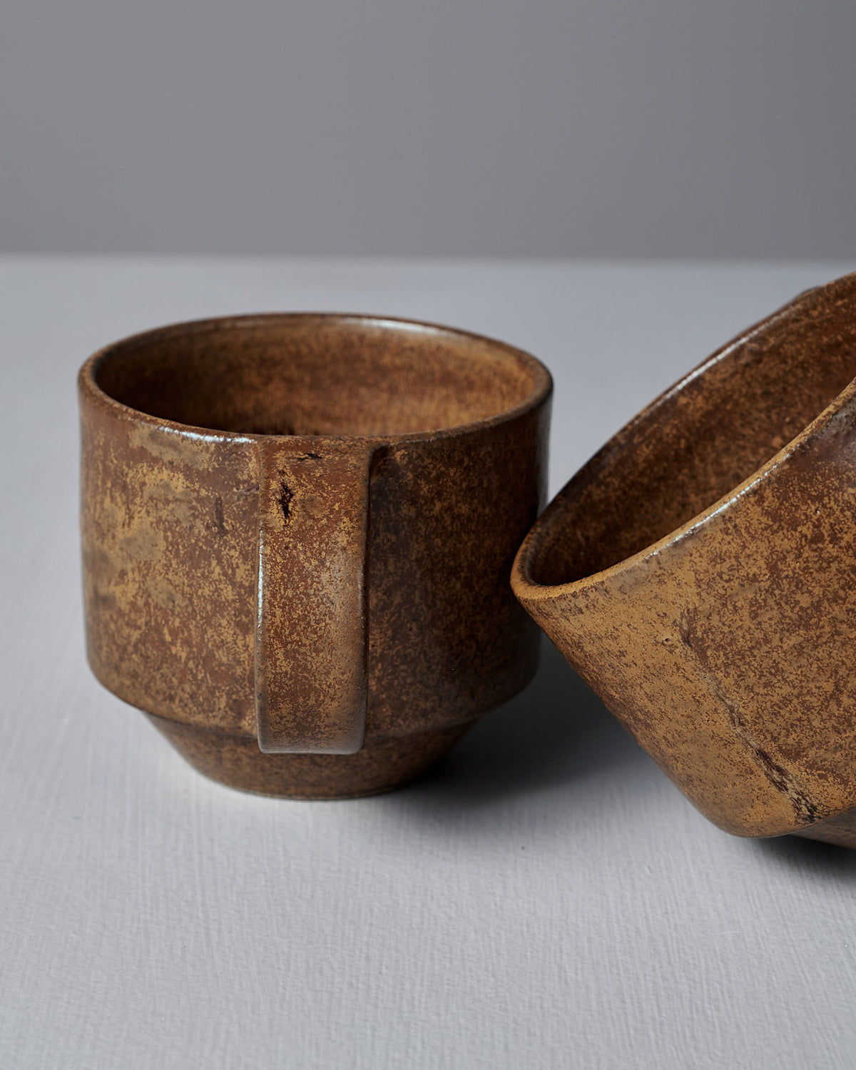Two Studio Foon Everyday Stacker Cups – Medium in terracotta, with a rustic, textured finish, are set on a light gray surface; one stands upright while the other lies on its side. Both are dishwasher safe and ideal for a handcrafted cup collection.