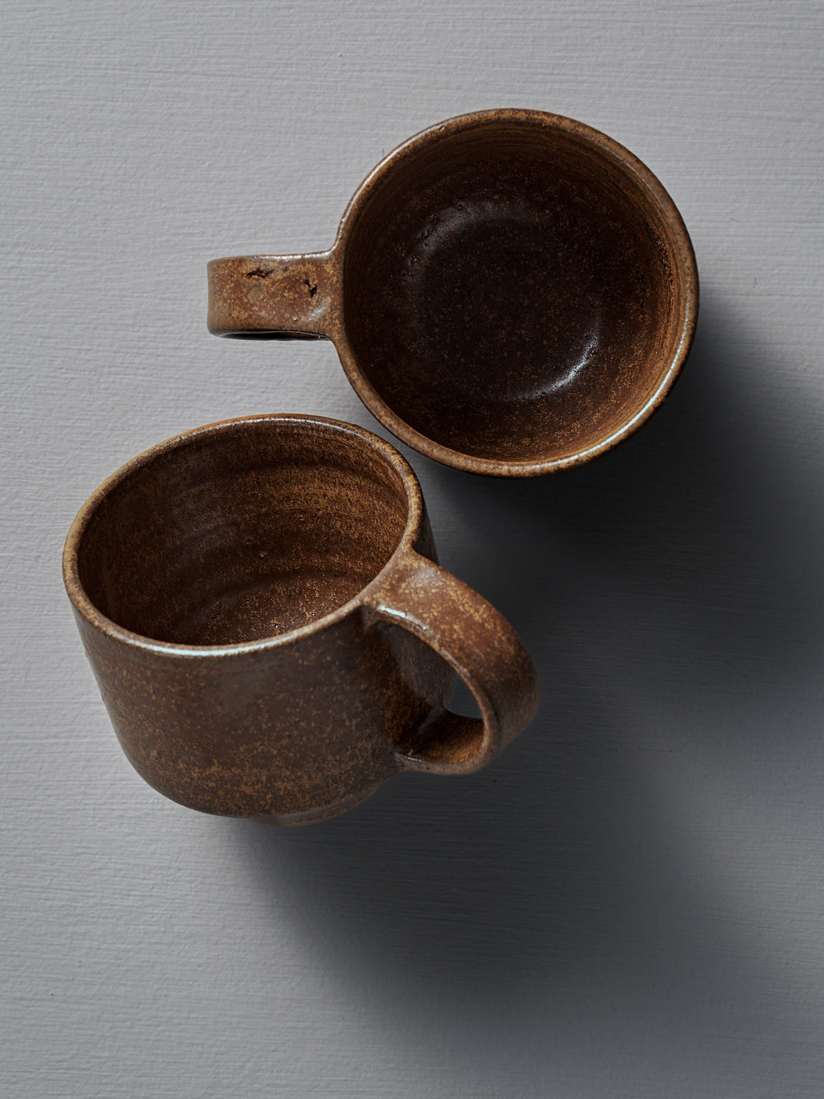 Two Studio Foon Everyday Stacker Cups – Medium in terracotta are displayed on a light gray textured surface, one upright and one sideways, both casting soft shadows.