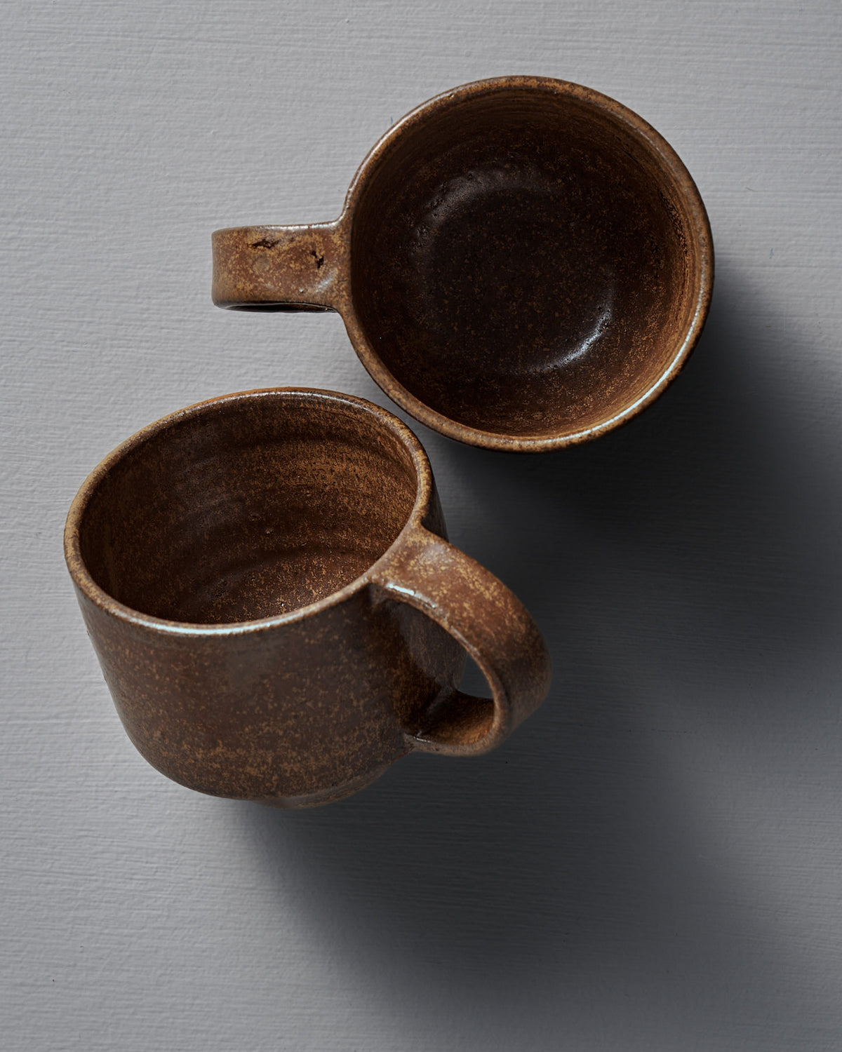 Two Studio Foon Everyday Stacker Cups – Medium in terracotta are displayed on a light gray textured surface, one upright and one sideways, both casting soft shadows.