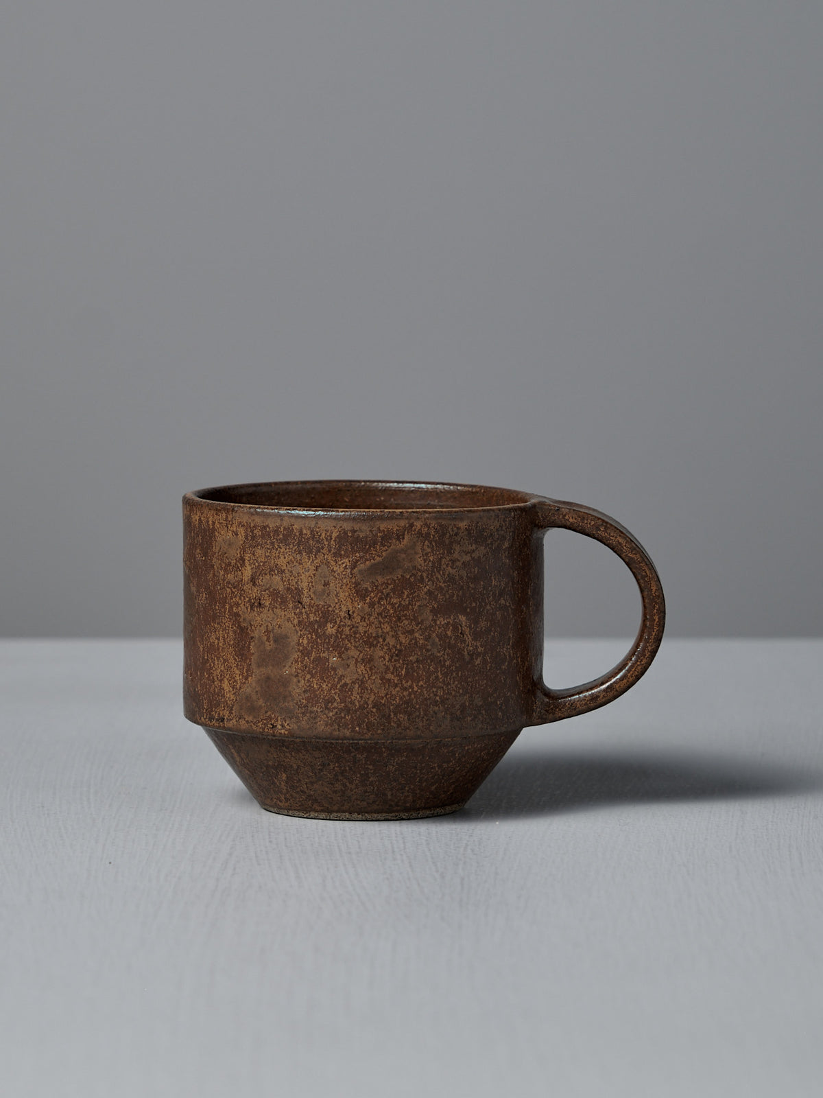 The Studio Foon Everyday Stacker Cup – Medium in terracotta, featuring a round handle, sits on a light gray surface against a plain gray background—ideal as your daily coffee cup.