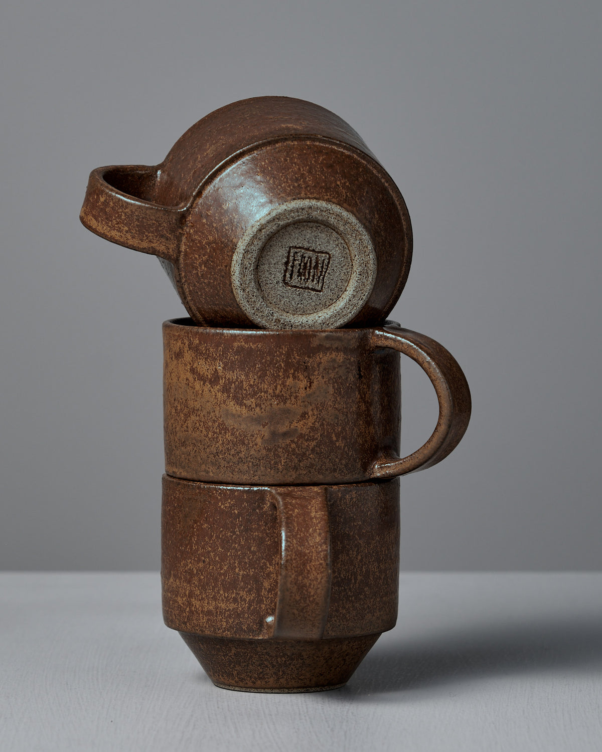 Three Studio Foon Everyday Stacker Cups – Medium in terracotta are stacked on a white surface against a gray background; the top cup rests sideways, highlighting its base with the maker’s mark.