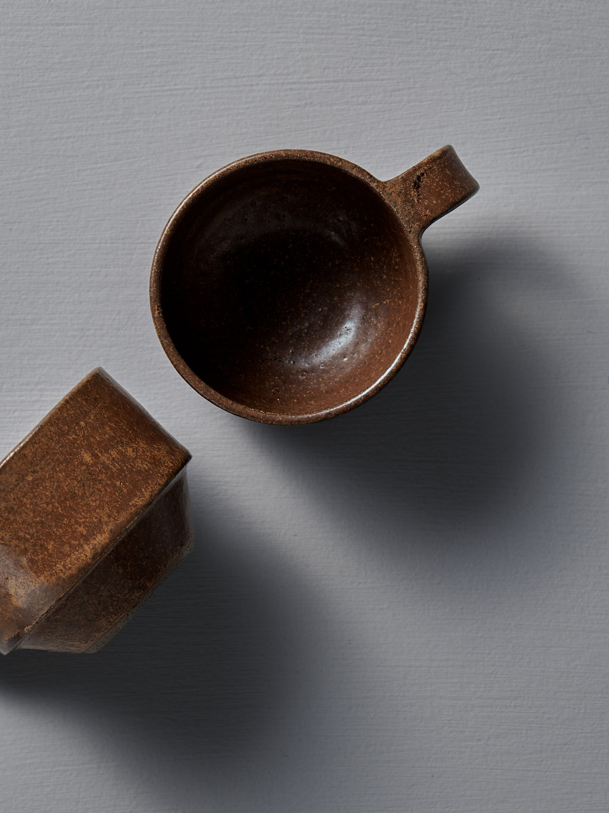 Two Studio Foon Everyday Stacker Cups – Small in Terracotta rest on a gray textured surface, one upright and the other tilted, casting soft shadows.